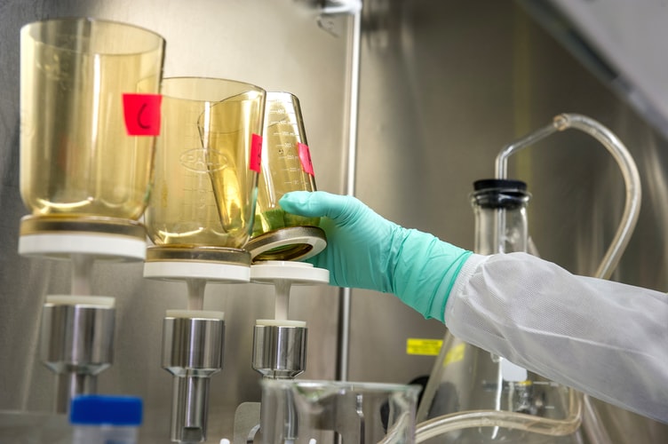 A gloved hand reaches for a beaker. There are three yellow-tinted beakers in a lab and an arm wearing a lab coat and green gloves picks up the third beaker.