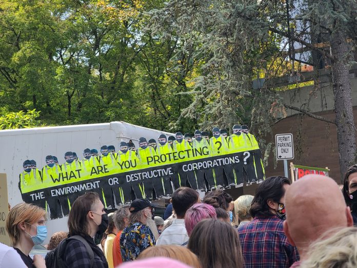 Background with board figures of police in uniform with words "what are you protecting?" on top and "the environment or destructive corporations?" at bottom. Foreground is people facing the right side.