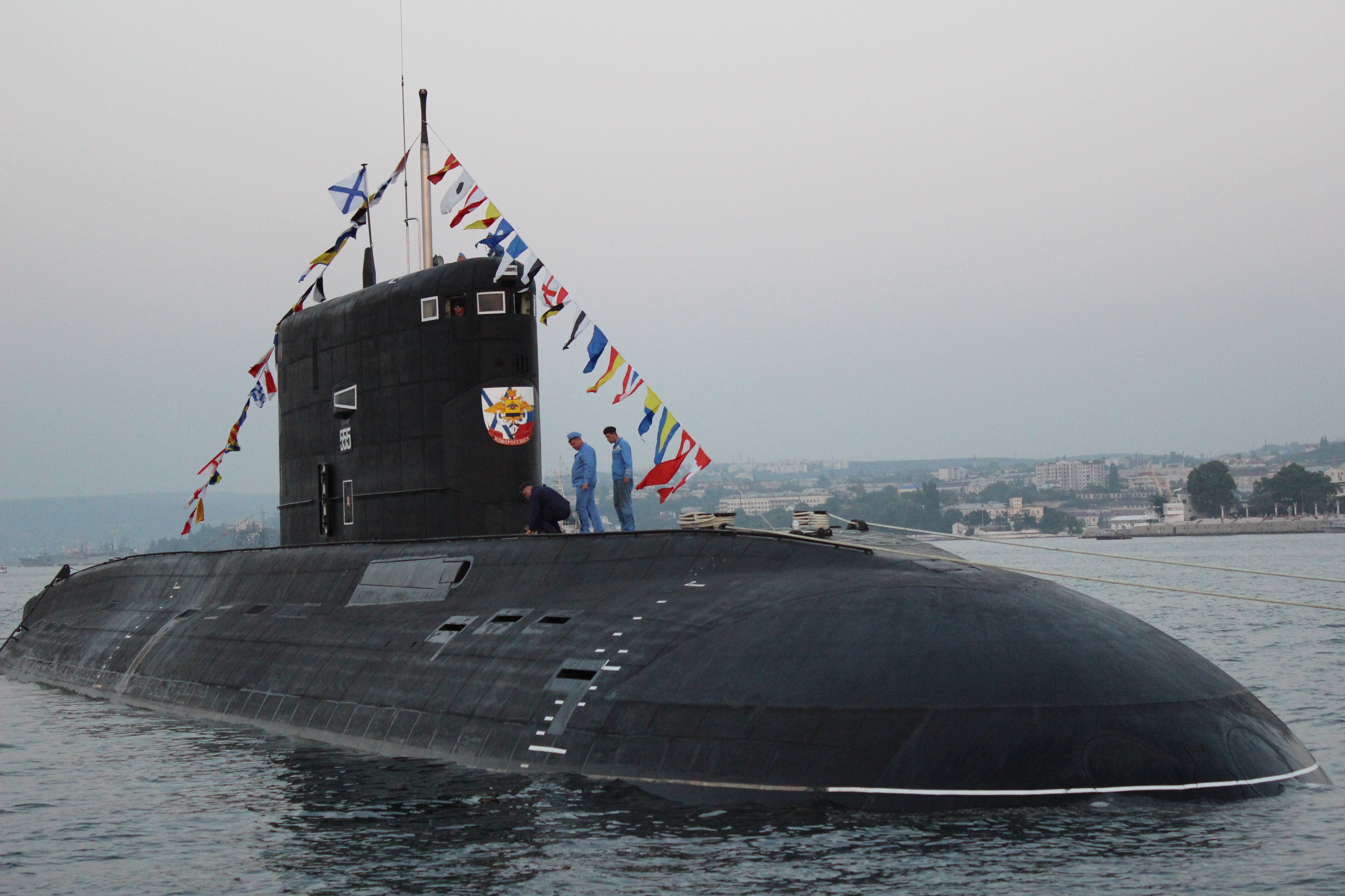 A partially submerged black submarine has two people standing on top. The submarine is near shore and the top has many countries' flags flown.