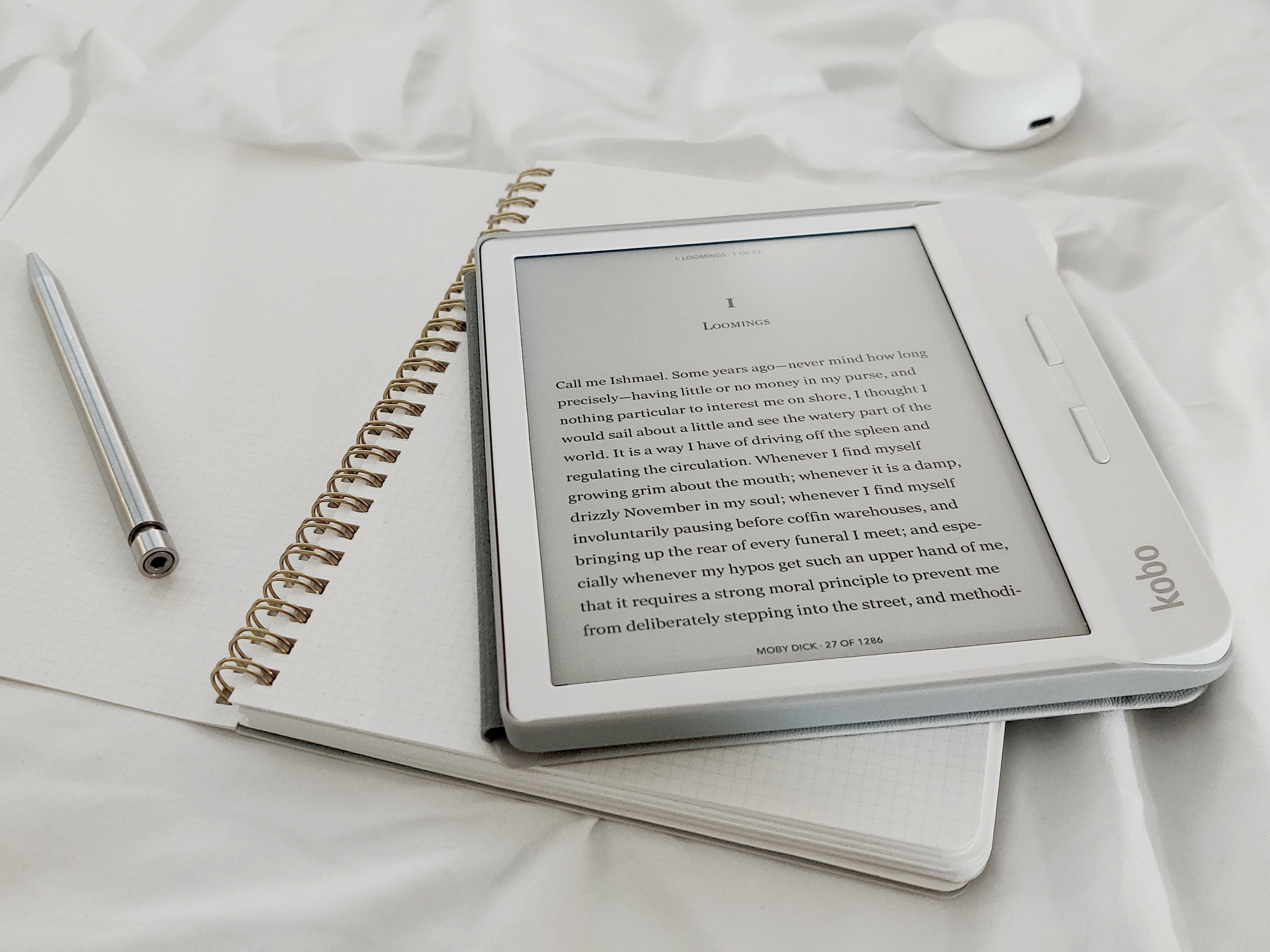 A white eReader with black text sits on top of an open and empty white journal. They are on a white sheet.