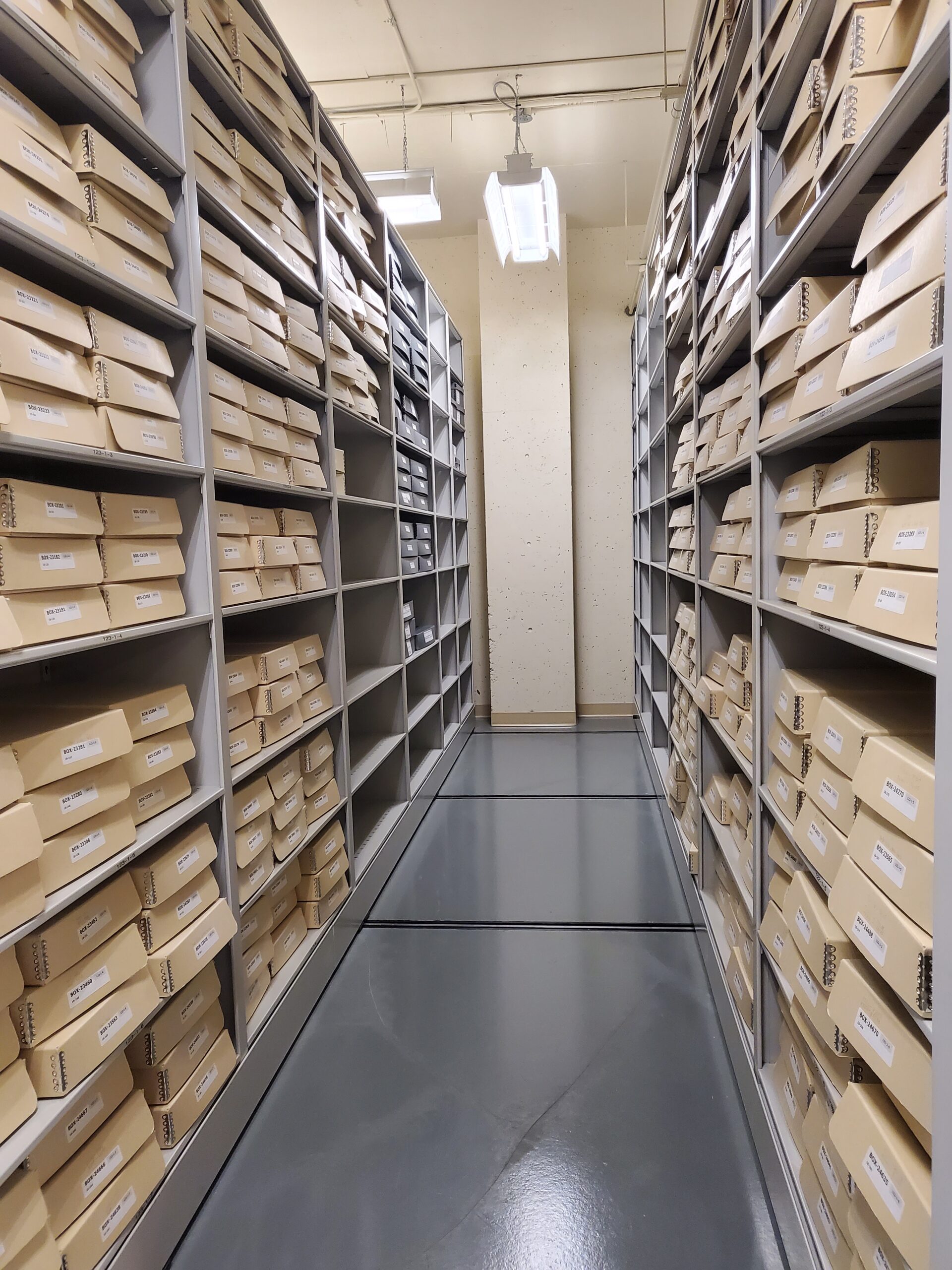 A hallway with shelves on either side holding beige archive materials. The floor is grey and is illuminated by fluorescent light. Overall, pretty bland.