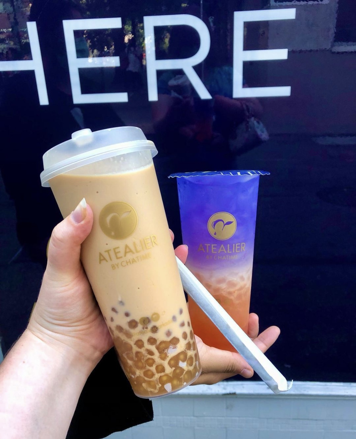 Two arms outstretched, holding large bubble tea cups: one milk tea with golden-coloured pearls and the other a shimmery purple drink with orange pearls