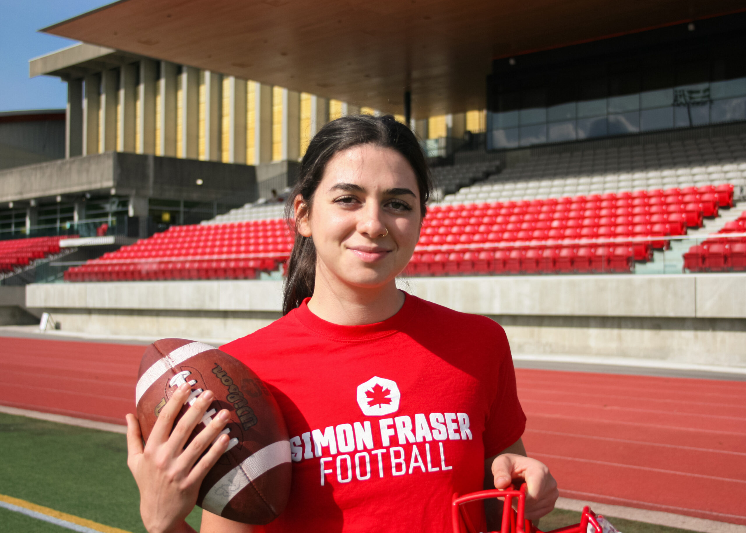 Kristie Elliot in a SFU athletics t-shirt holding a football. Background is SFU stadium