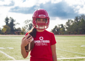 Kristie Elliot in SFU athletics t-shirt, holding a football on her left while wearing a helmet 