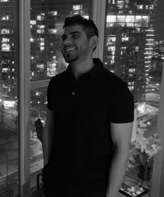 Black and white image of Bains, a South Asian man, standing in front of a floor to ceiling window with a cityscape view