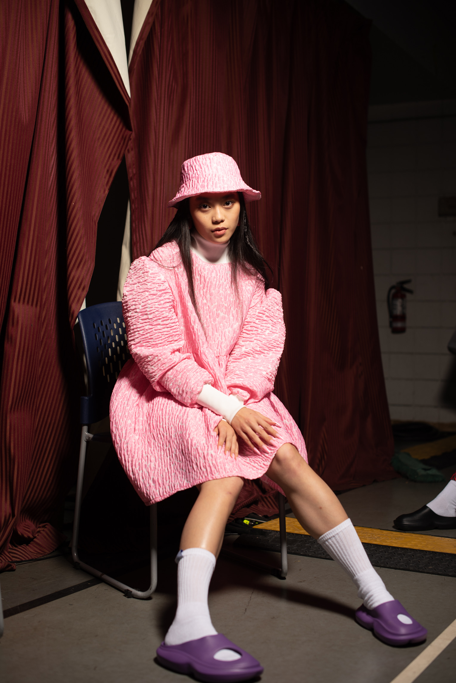 A model, wearing a bubbly pink dress and matching bucket hat, sits backstage with her arms in her lap. She stares directly at the camera