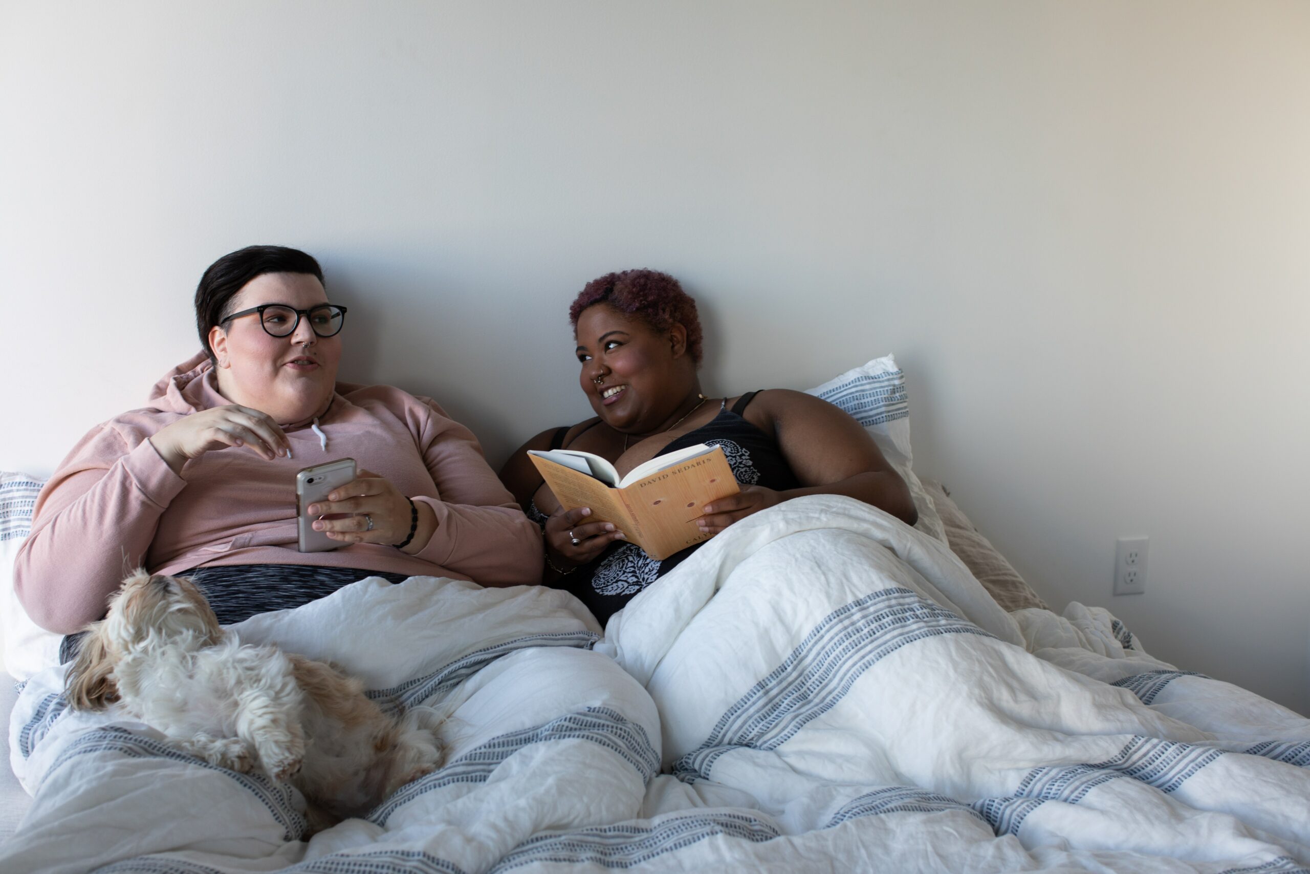 Two people and a dog are sitting on bed. They are smiling, laughing and talking to each other. One is holding a book and one has a phone.