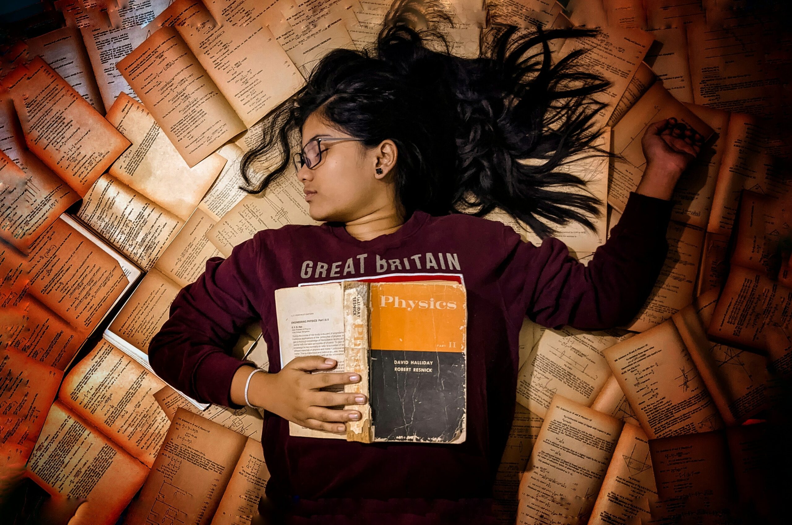 A person with long black hair asleep in a pile of books with one open across her chest