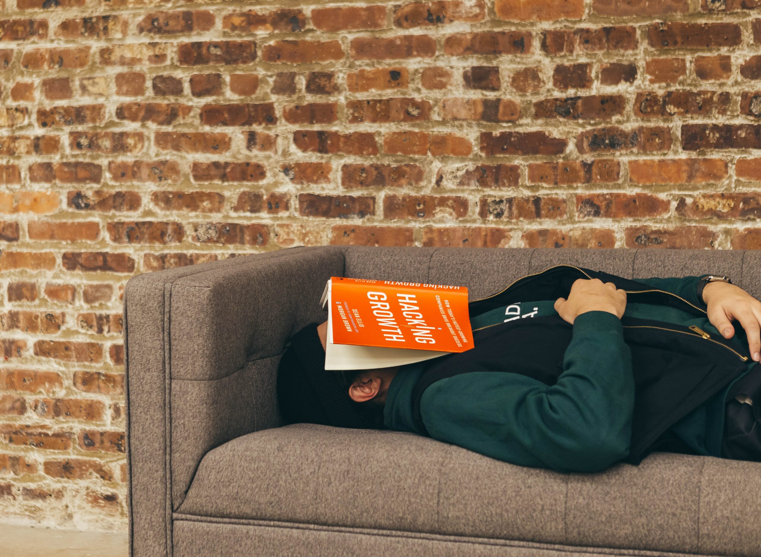 person sleeping on brown couch in brick room with red book spread over his face
