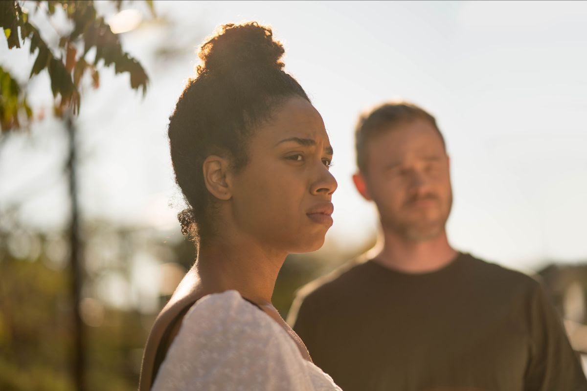Side profile of a Black woman, sunlight streaming behind her. Also in the background is the woman's partner, but he's not in focus