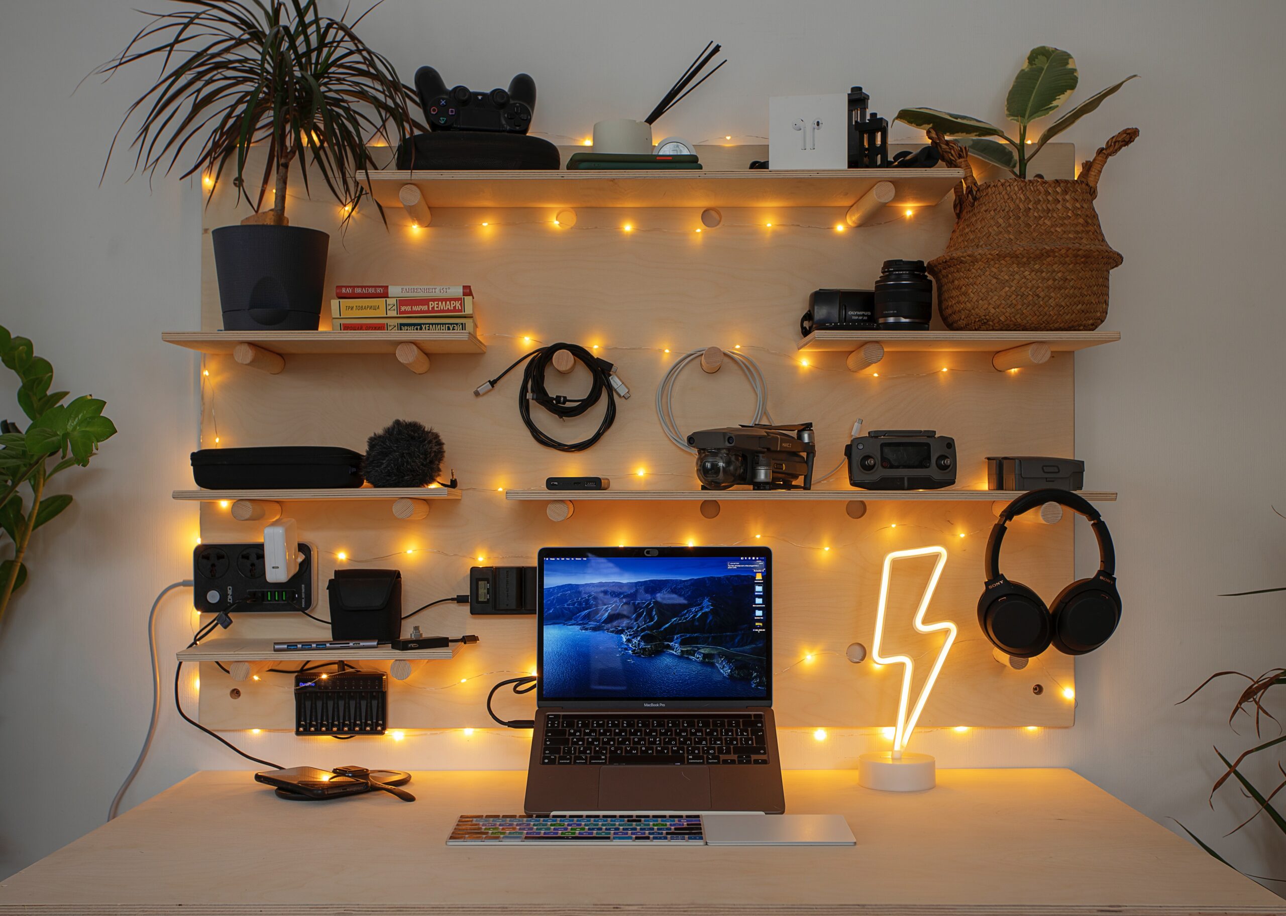 A pale wooden work station. On the desk: a keyboard, laptop, router, and lightning bolt lamp. On the shelves above: plants, books, chargers, headphones, a camera lens, and video game console. Fairy lights hang in between the shelves.