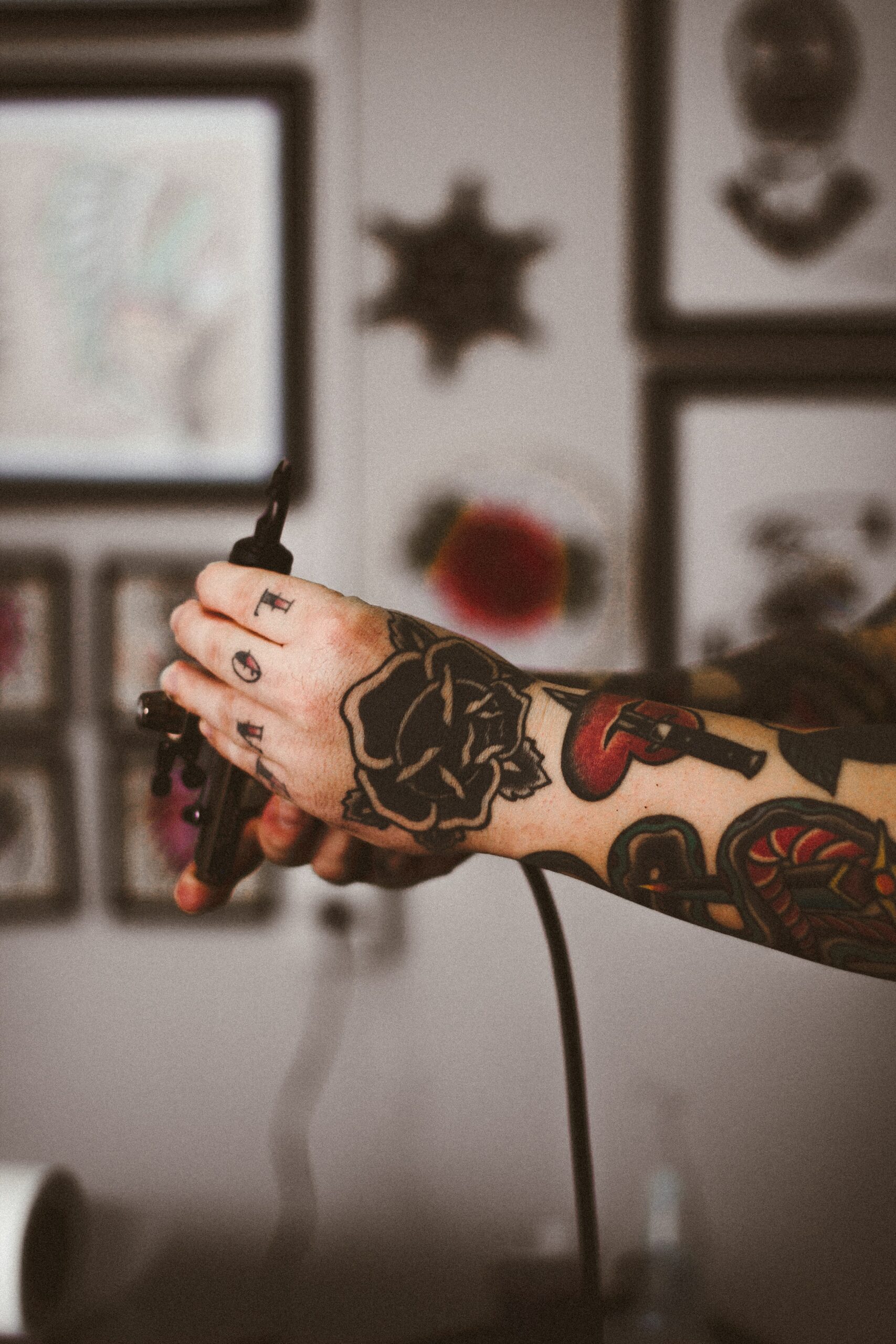 Inside a tattoo parlour, white background wall with framed designs blurred as image focuses on outstretched arms, covered in red and black tattoos (a heart with a sword going through it, flowers, etc.). One hand grasps a black needle