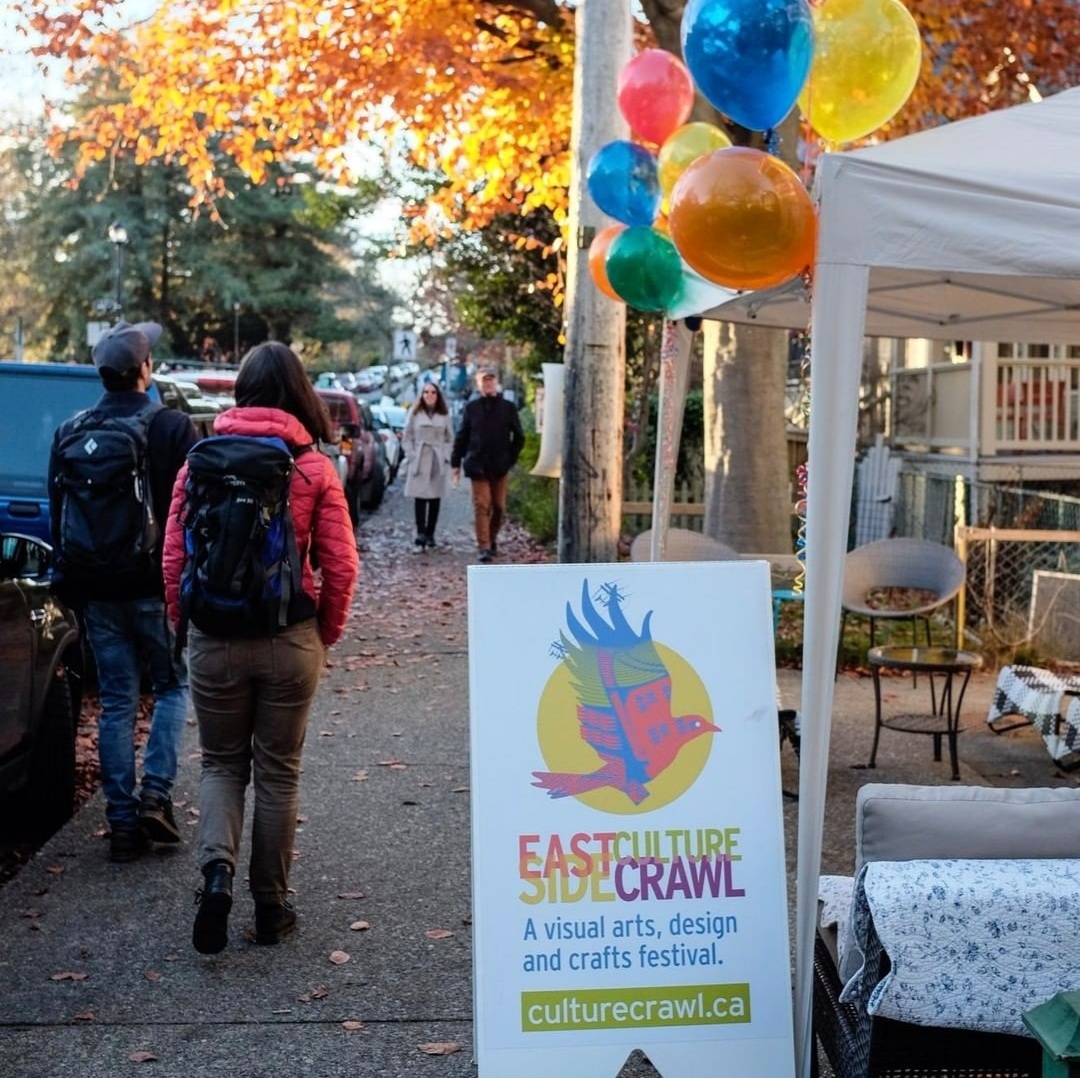 A sunny fall day on an East Vancouver residential street. People walk to and from a tent on the right with colourful balloons, streamers, and an Eastside Culture Crawl sandwich board.