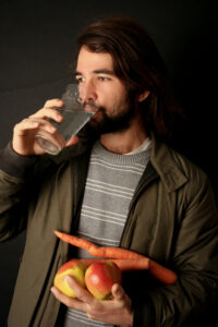 Jacob drinking a glass of water. They hold an armful of vegetables and wear a jacket