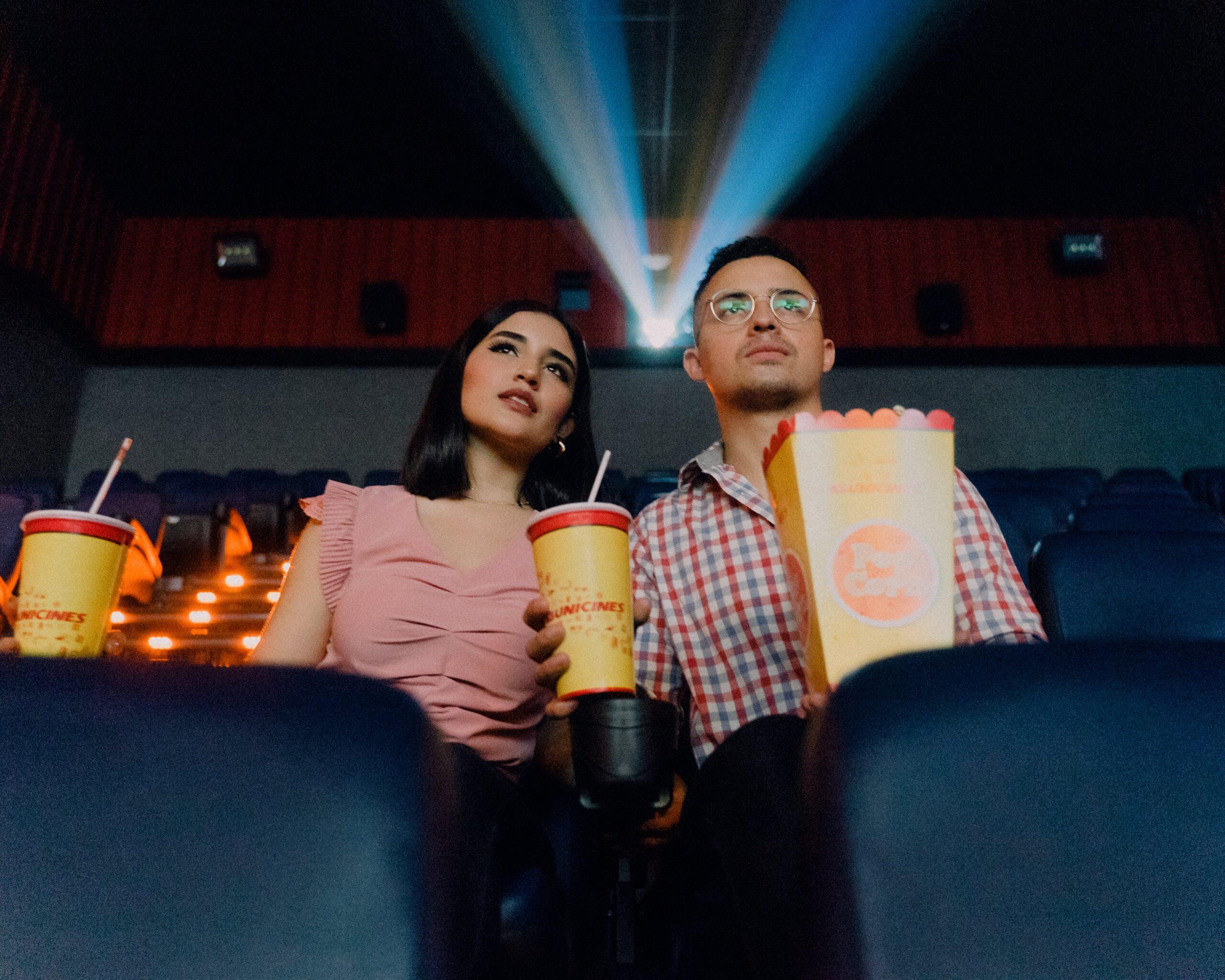 A young couple are at the theatre. The projector light passes overhead, as the two stare over the camera. A drink is between them, and a bag of popcorn and another drink are at their sides. They look slightly disconcerted about something happening on screen.