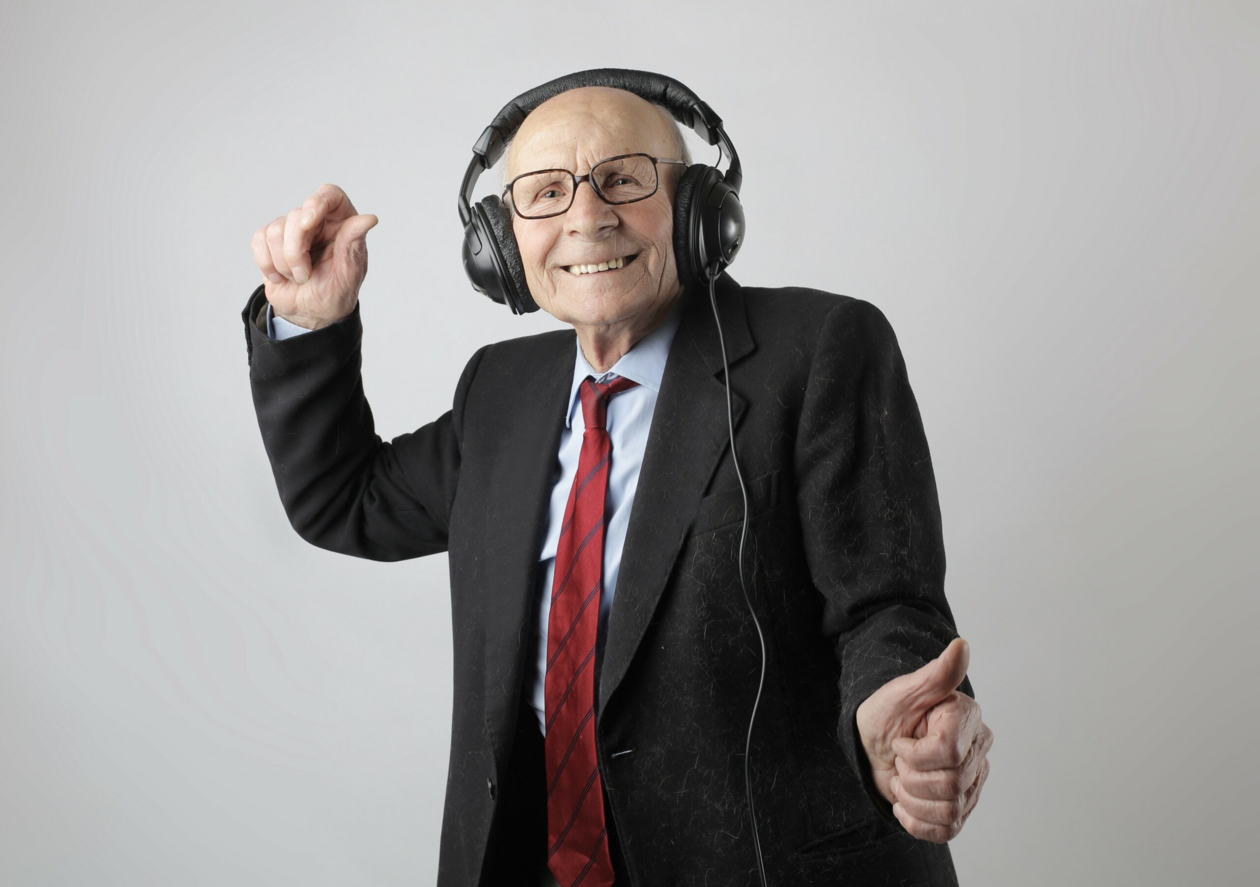 An old man in a suit is dancing. He is wearing large headphones, and is smiling at the camera.