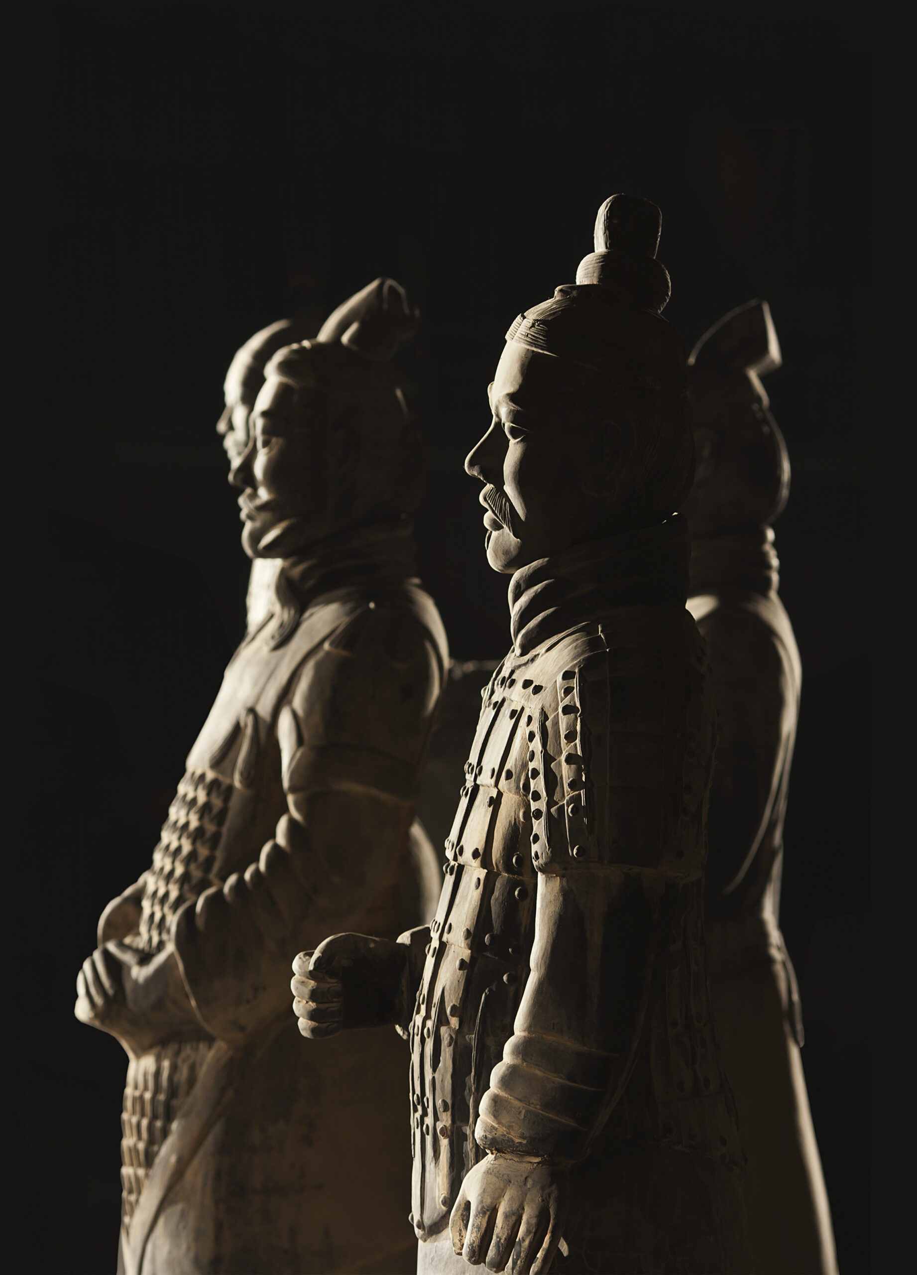 Four grey stone statues of Sun Tzu standing beside each other all facing towards the left. The lighting is dim and dramatic with a black background behind the statues.