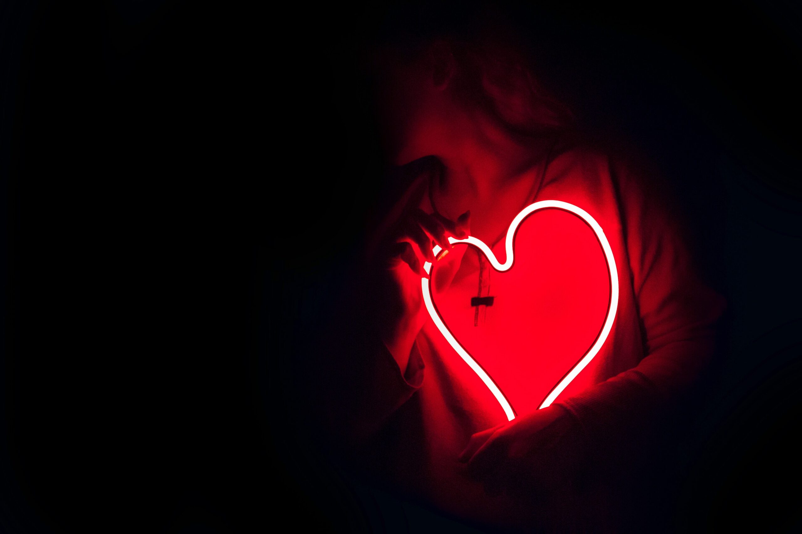 A person is holding a red heart-shaped neon light. The neon is the only light in the photo, and the person is mostly obscured by the darkness.