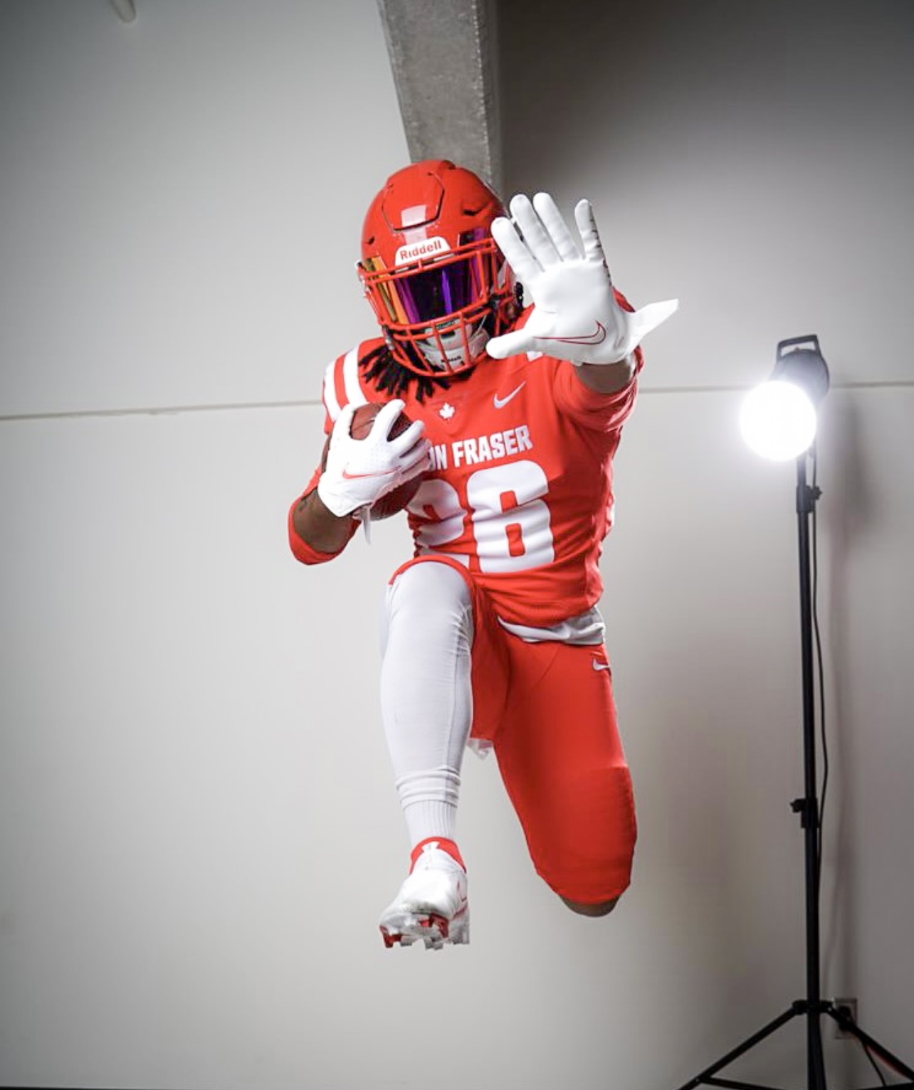 A photo of SFU football player Mason Glover in his gear jumping in the air with a football in one hand and the other being held up to the camera.