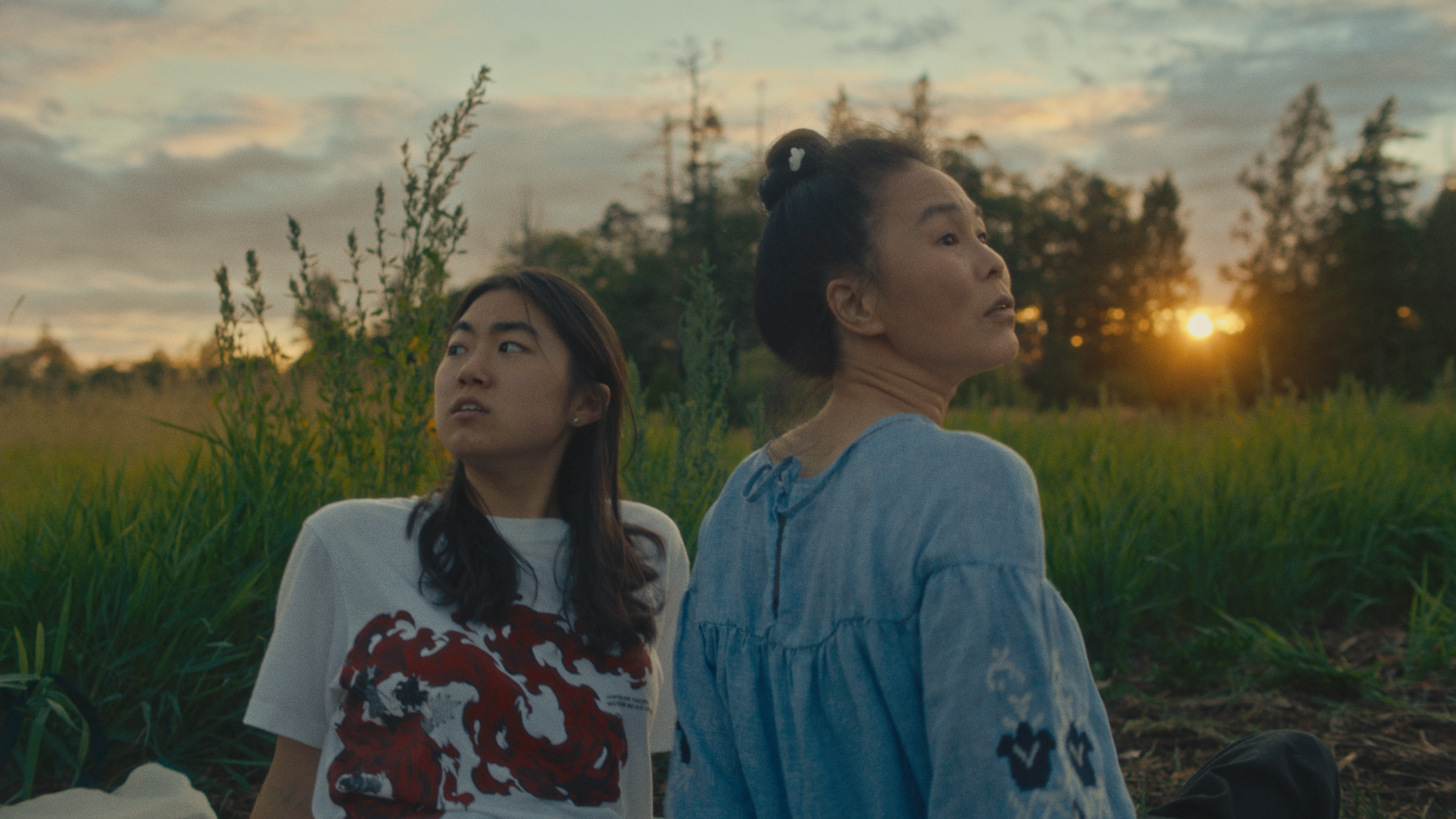 Two Asian women, a granddaughter and grandmother, sit in a green field with the sun setting behind them