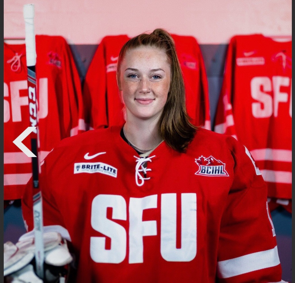 Photo of SFU goaltender Kayla Munro smiling for the camera in her gear in front of three SFU jerseys in the background.