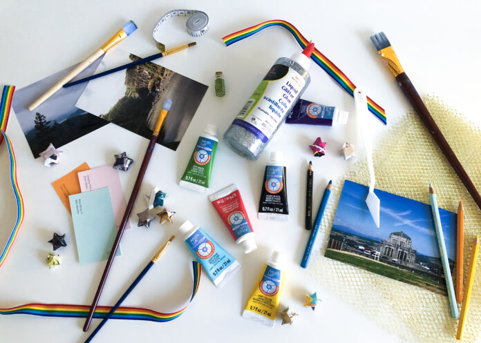 Flatlay photo of a white table full of art supplies — like acrylic paint, pencil crayons, and cardstock — in a variety of colours.