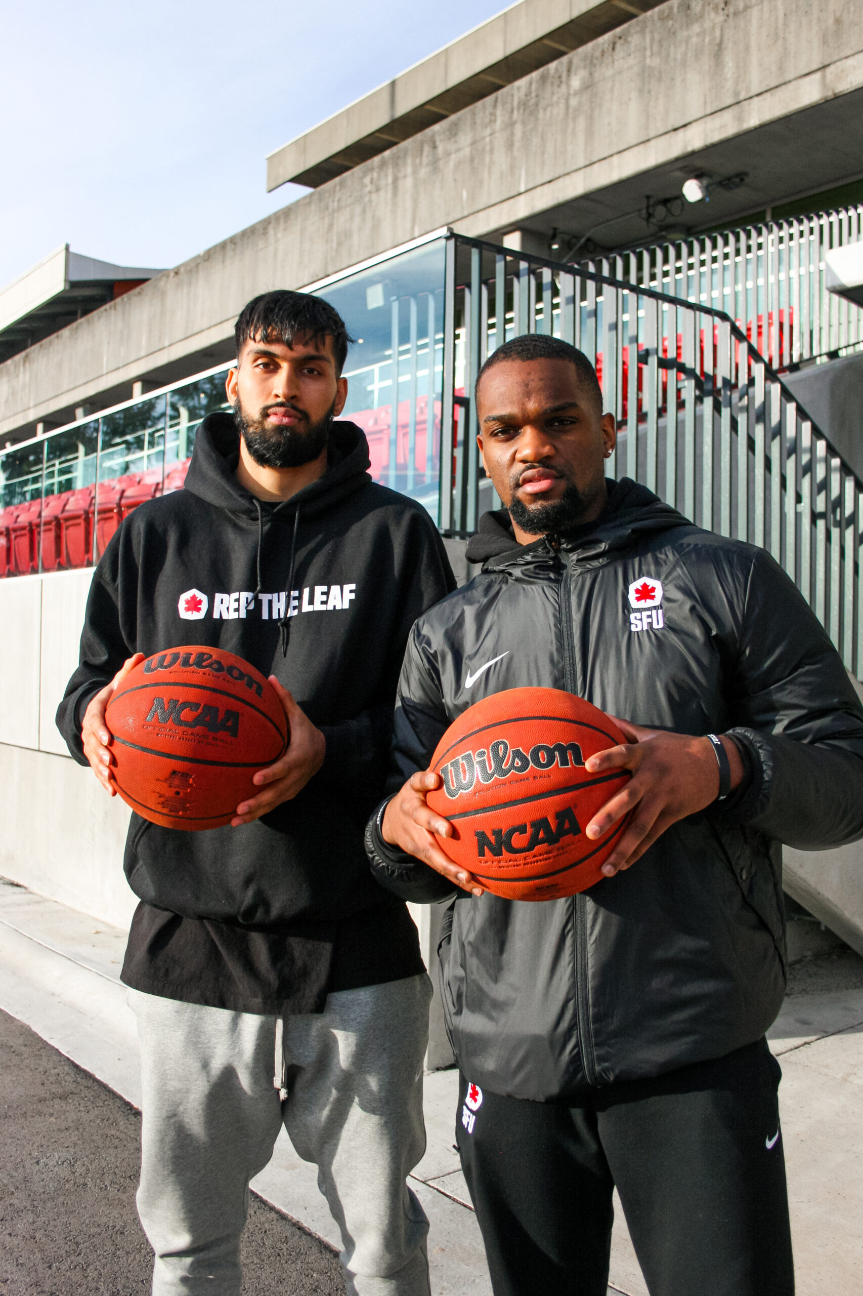 SFU basketball players Jas Singh and Wilfried Balata stand in front of the camera posing with a basketball in their hands.