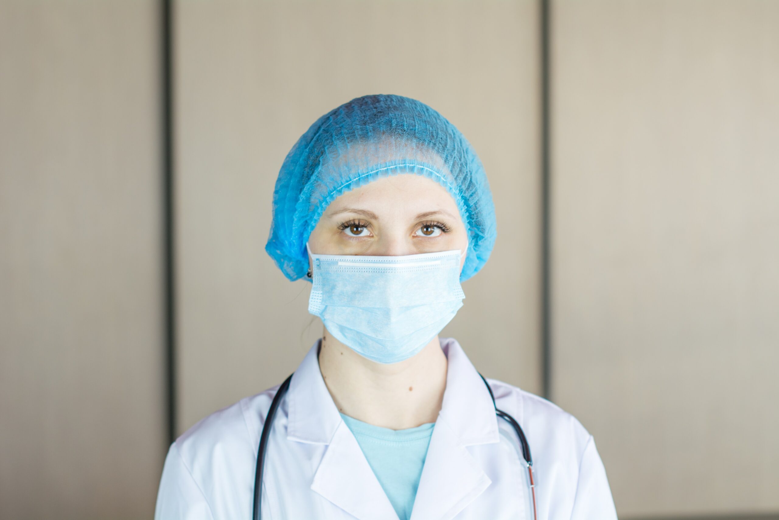 A woman is dressed in medical equipment, wearing a face mask and a head covering. A stethoscope is draped over her shoulders, and she is looking at the camera unflinchingly, perhaps with some exhaustion.