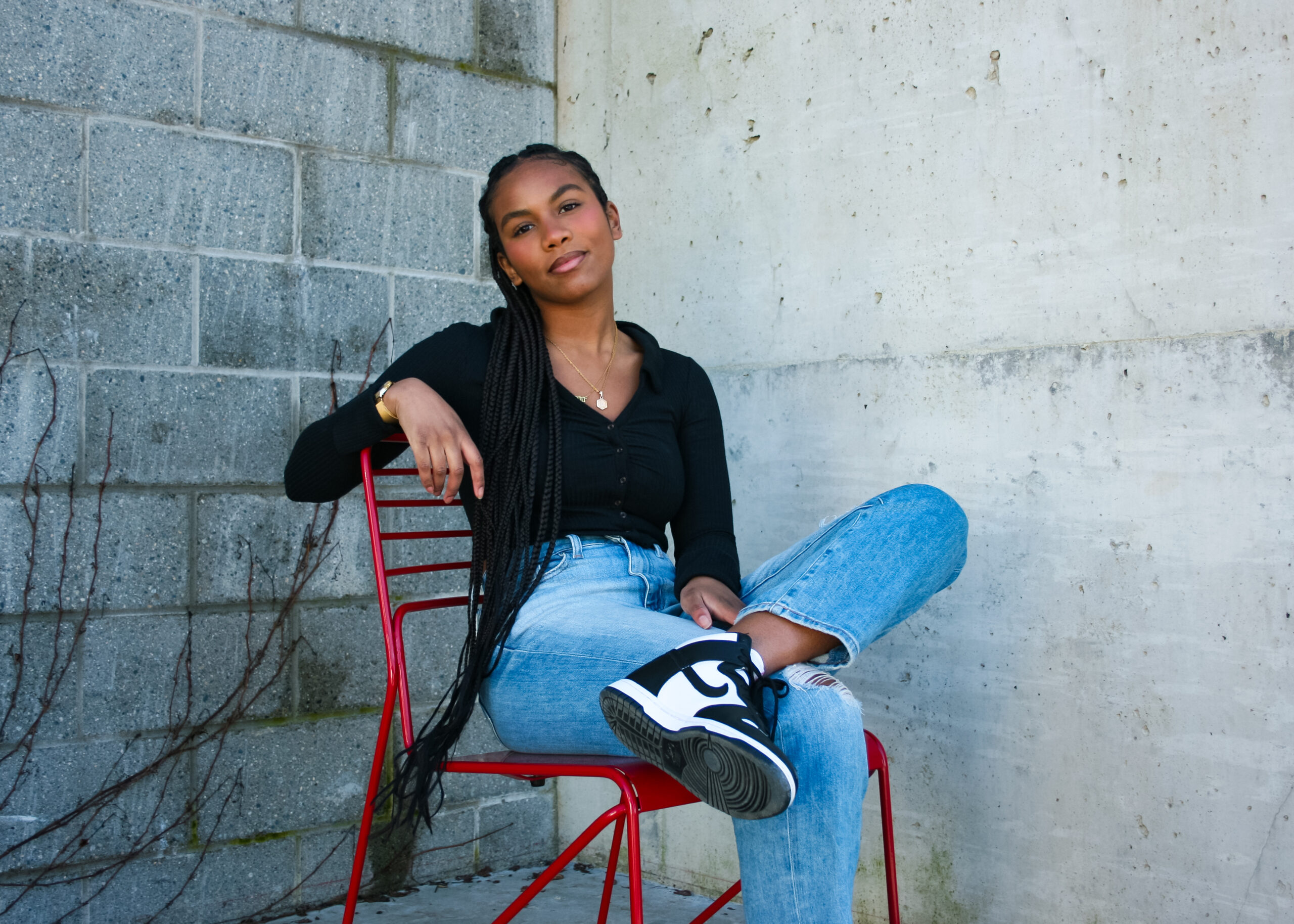 Linda, a Black-Filipinx woman, sits in a red chair, legs crossed, with brick and concrete walls behind her. One arm is draped over the back of the chair as she stares straight at the camera.