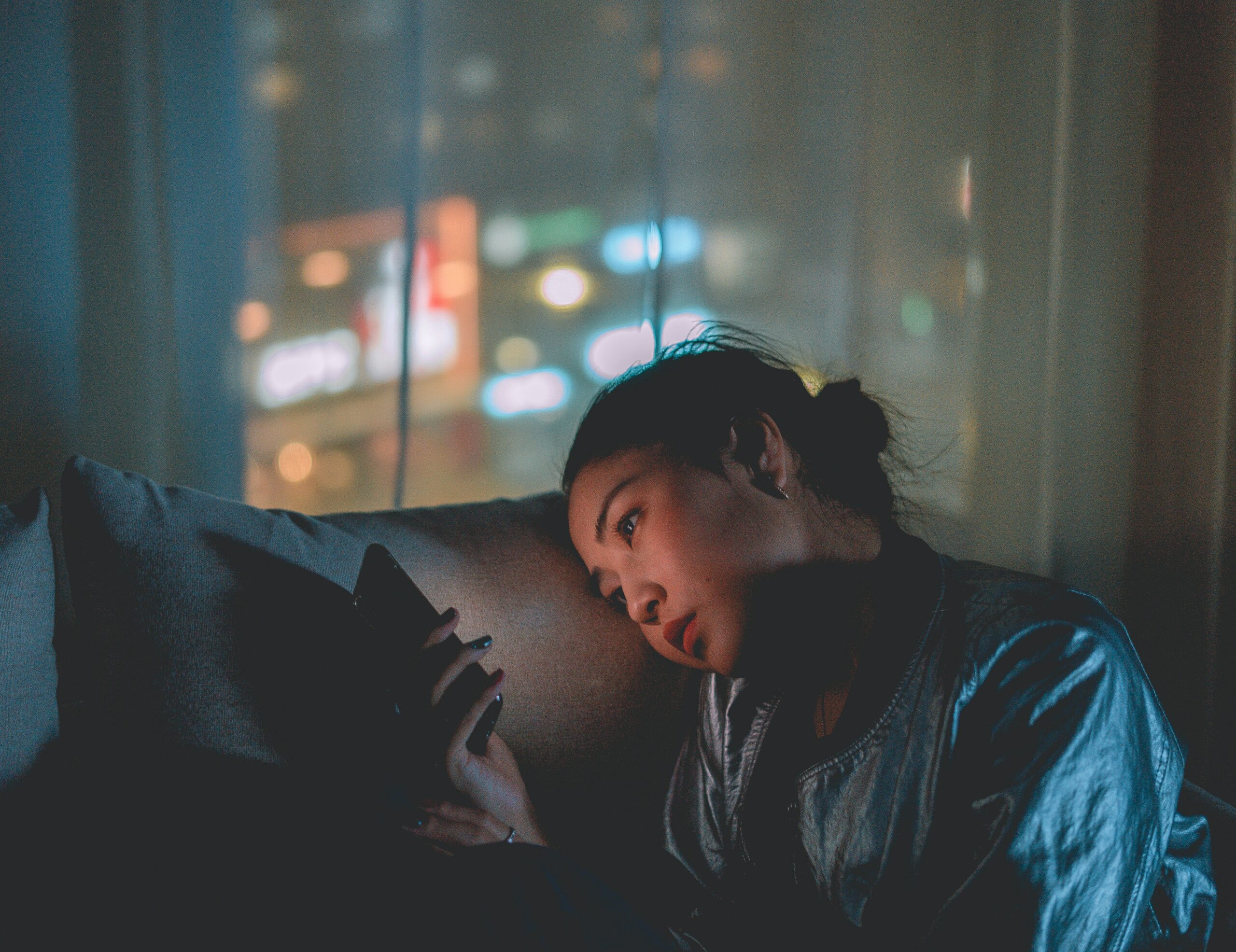 A woman is looking wistfully at her phone screen. The room is dimly-lit, and curtains behind her filter out some of the bright lights of the city. The mood of the photograph is very isolating.