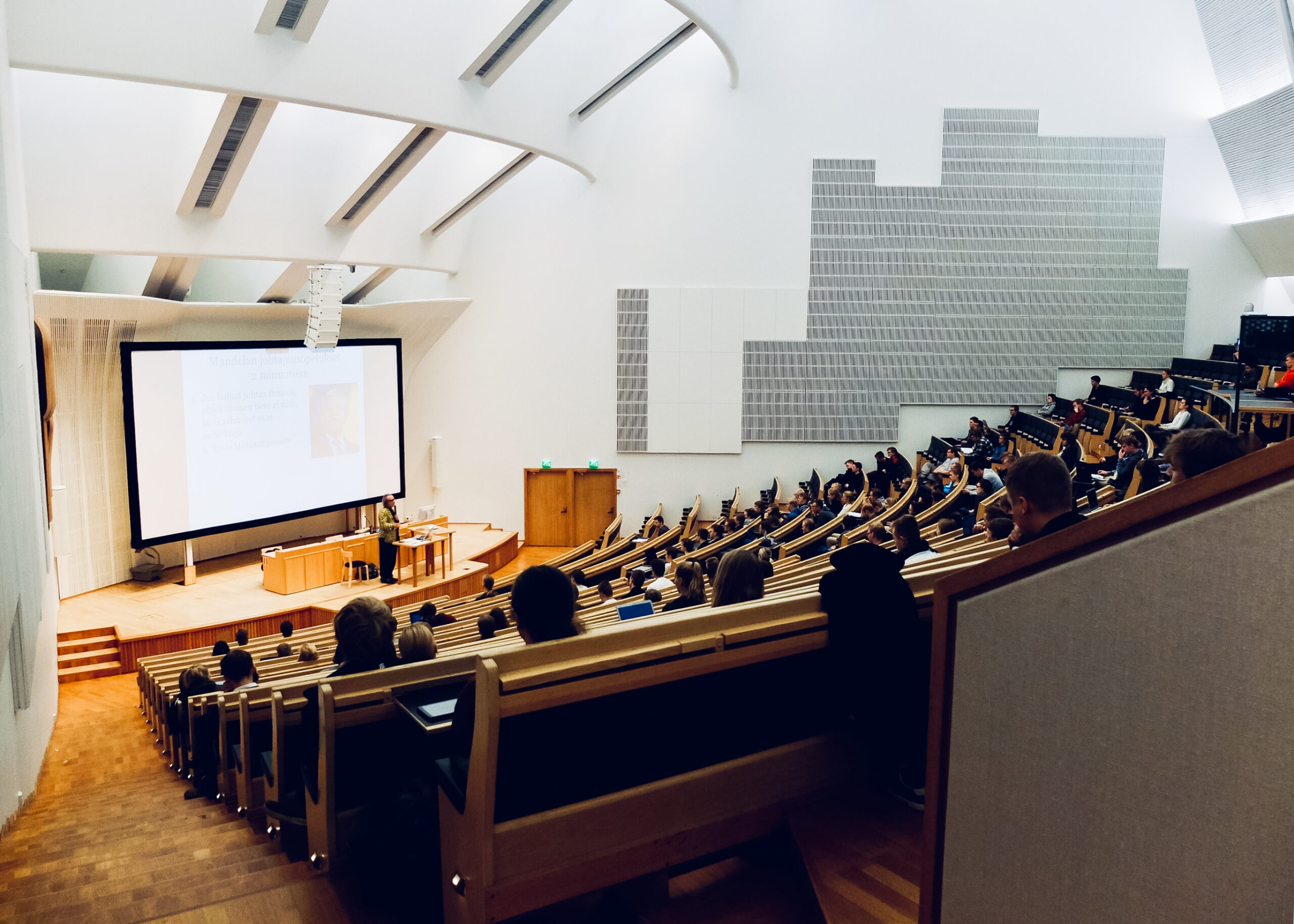 A professor on the right is pointing at a very large screen and lecturing to a lecture hall full of students on the right side of the photo. They are at wooden tables that curve inwards to face the professor at the front of the room.