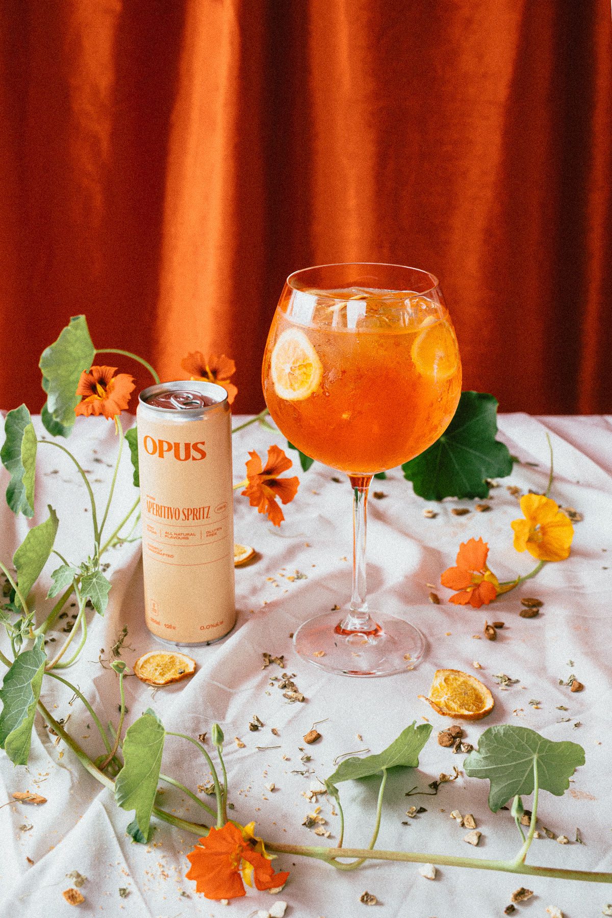 A white tablecloth covered in flowers, dried fruit, and spices. Standing upright is a glass filled with aperitivo spritz, next to an opened OPUS can