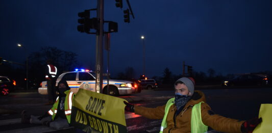 Two protestors holding a banner that says “Save Old Growth” with a police officer standing in the background, along with a police car.