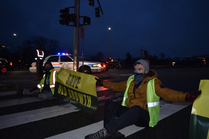 Two protestors holding a banner that says “Save Old Growth” with a police officer standing in the background, along with a police car.