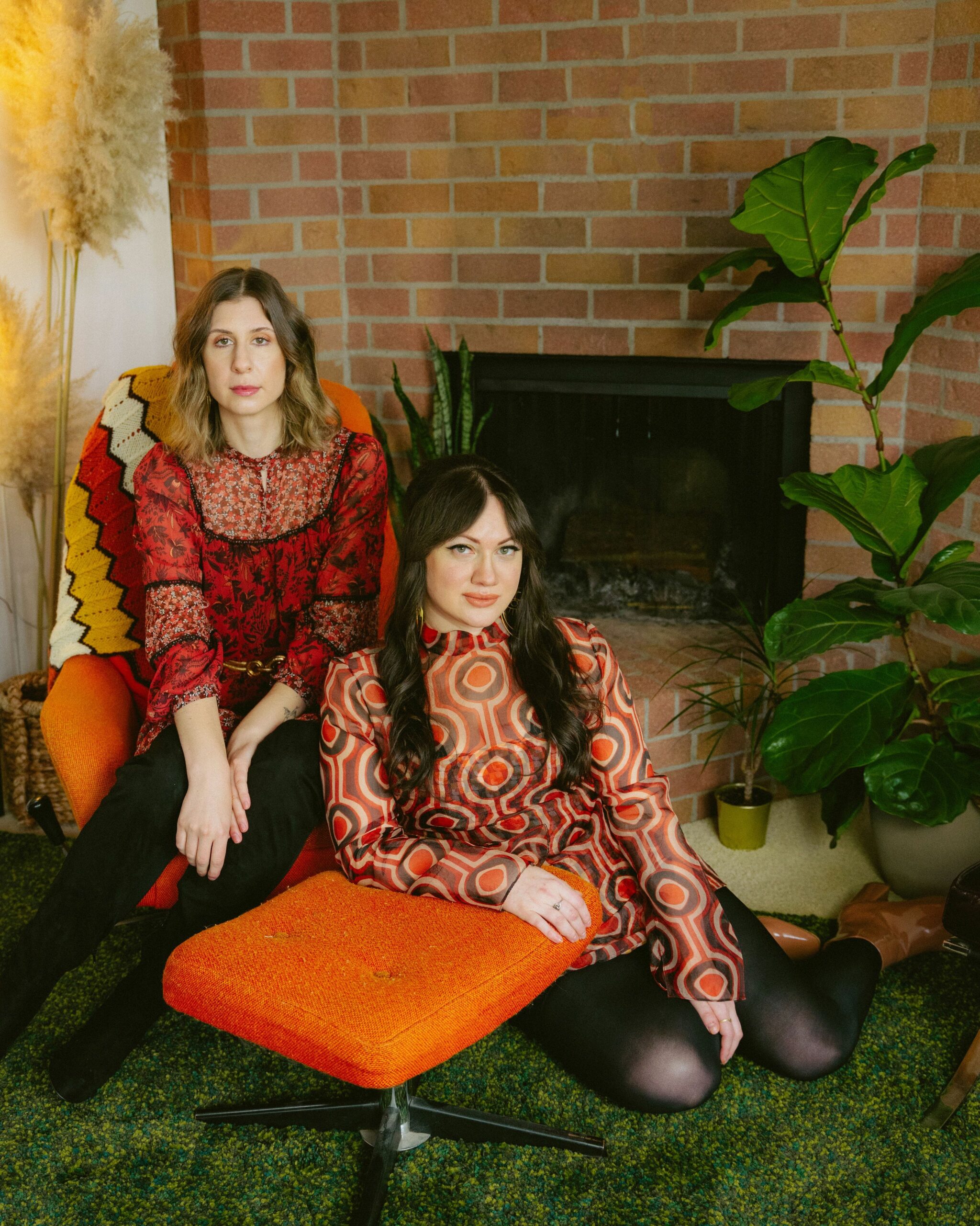 Emmalee (left) and Becky (right) sit on or by fuzzy orange furniture, set in front of a brick fireplace and flanked by potted plants