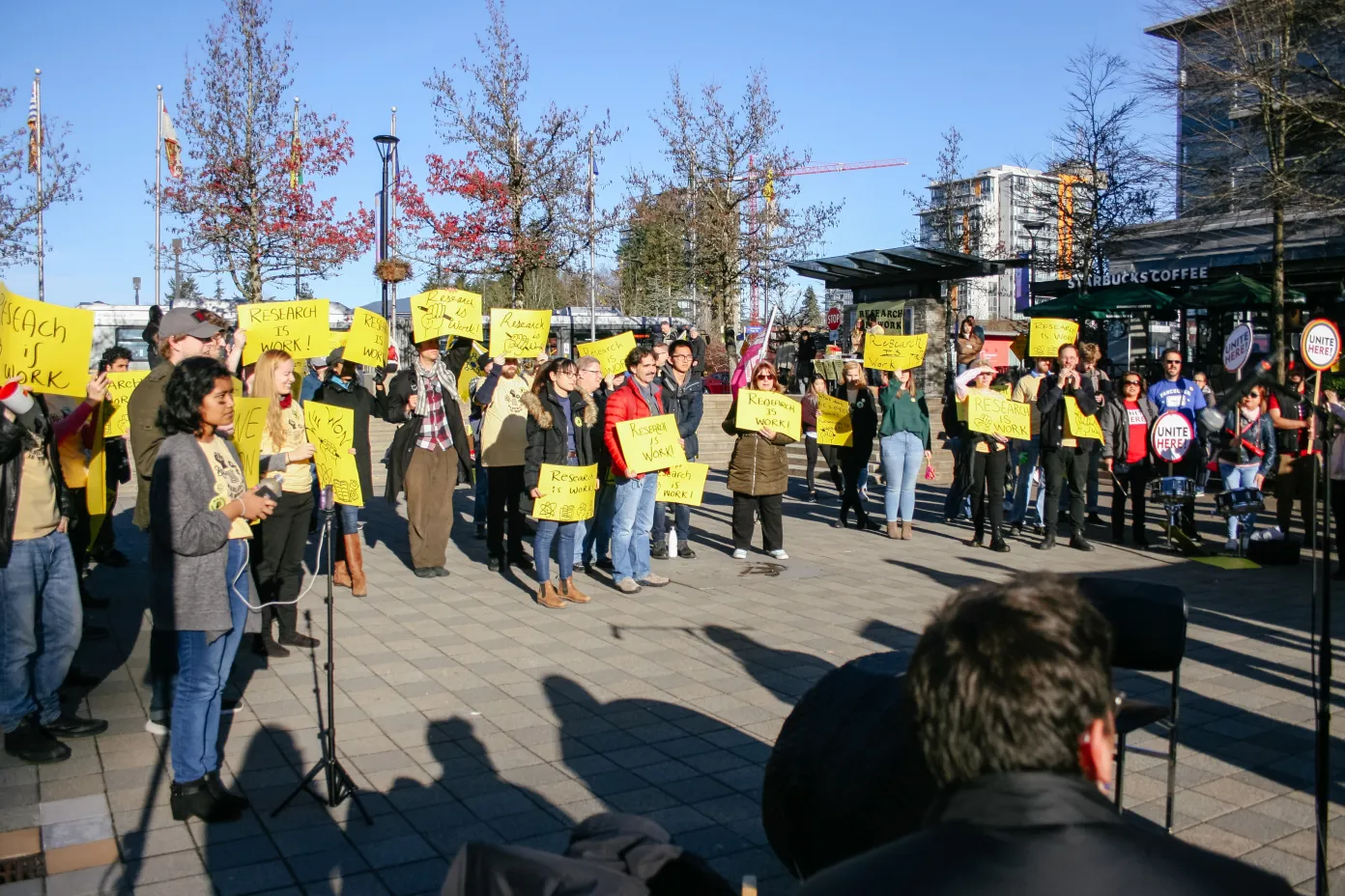 A group of people gathered around Cornerstone at SFU Burnaby campus holding yellow signs that say “Research is work”