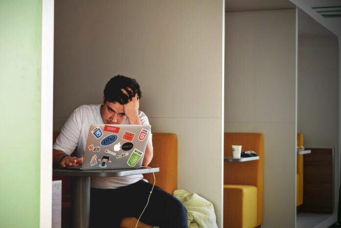 A student is in a booth, running their hand over their forehead. A laptop is open in front of them, decorated with stickers. They look stressed about broad circumstance, which is not helped by whatever they’re seeing on-screen.