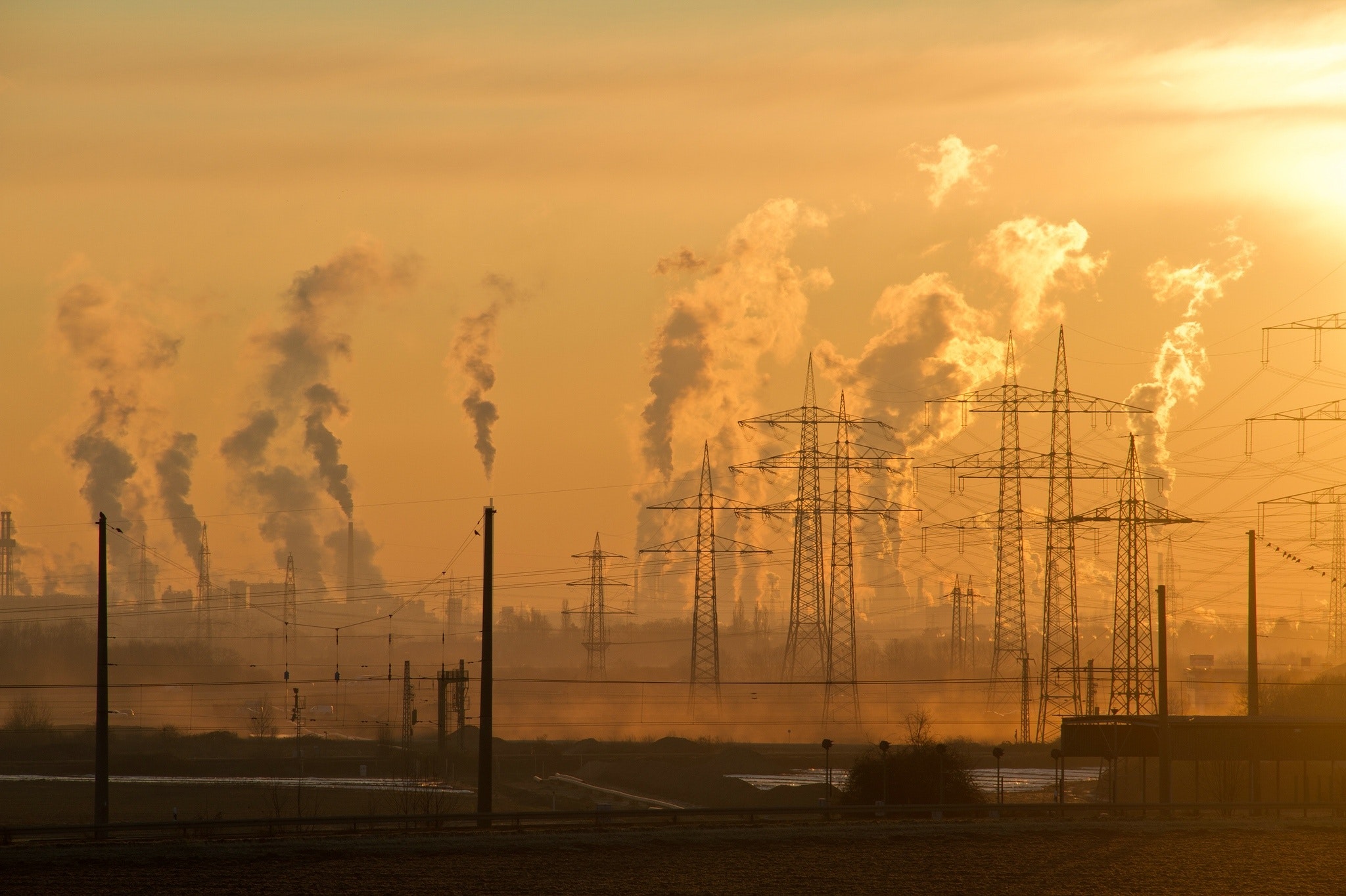 A power generation facility. Electric lines fill the photo, and pillars of something cloudy comprise the background. The day looks hazy, hot, and pervasively dirty.