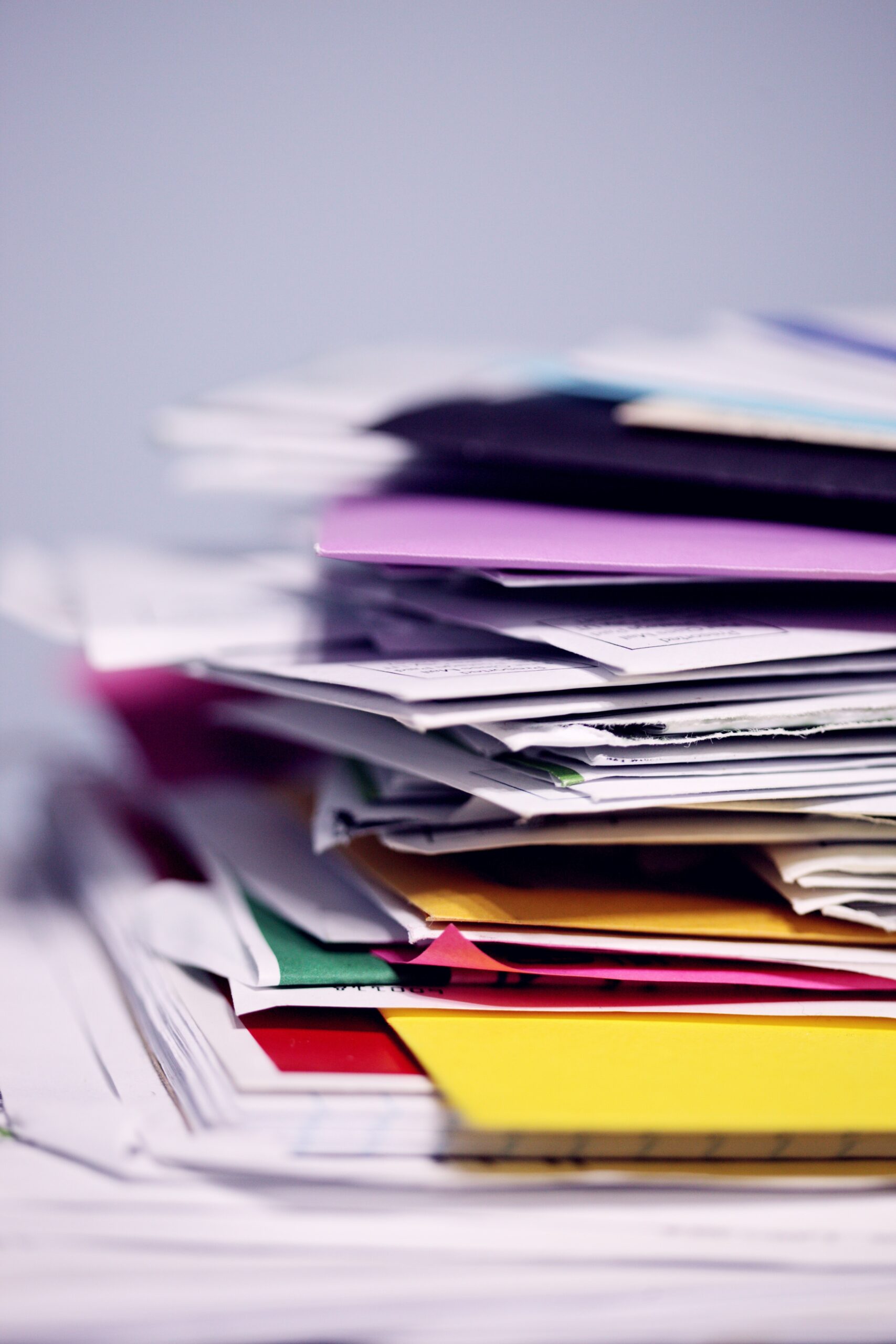 A pile of folders and papers are stacked up for the entirety of the photo. Some of the papers are folded, and the pile looks in disarray. The folders are brightly coloured and stand out.