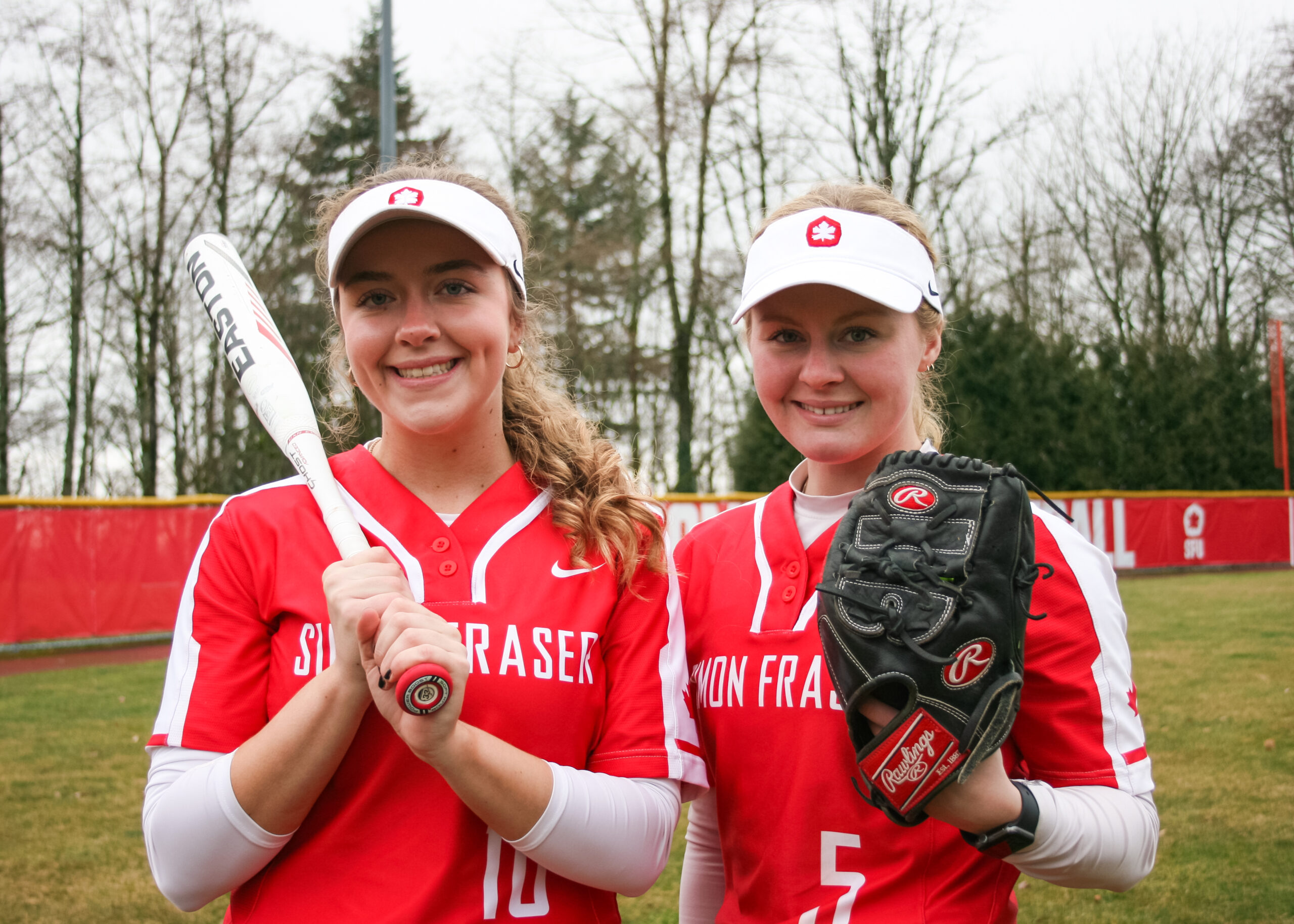 SFU softball players Rebecca Kirkpatrick and Lauren Schwartz pose with a bat and glove in uniform on the field.