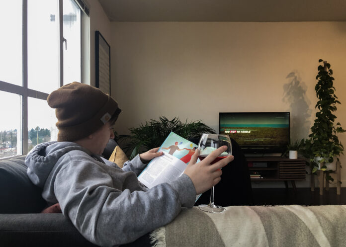 A person is reclining on a couch. The photo is taken over their shoulder, and we can see that they have a book open, a glass of wine, and a TV playing in the background.