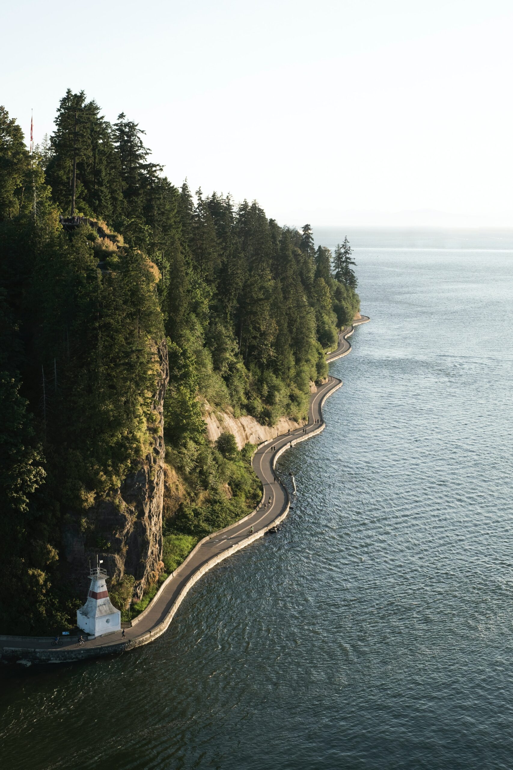 A photo of the Stanley Park seawall. A pedestrian path separates the image into ocean and forest, and a small lighthouse is anchored in the foreground. The sky is slightly overcast, and almost blends into the sea.