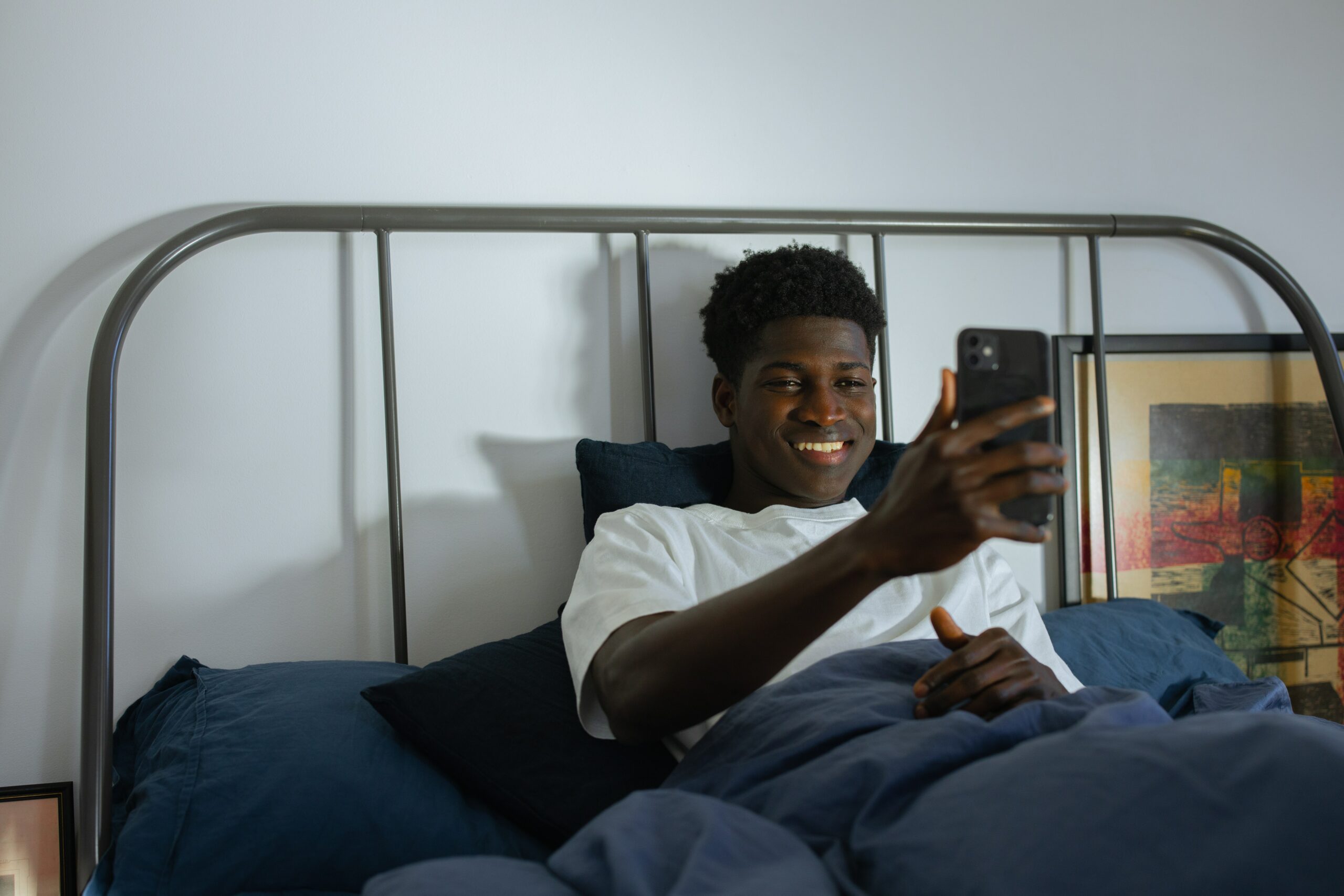 A guy is lying in bed, holding his phone like he’s on a video call, and smiling.