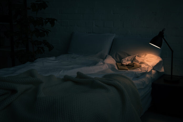 A dimly lit room with a bed and a book open on top of the bed. The only source of light is coming from a small lamp on a bedside table.