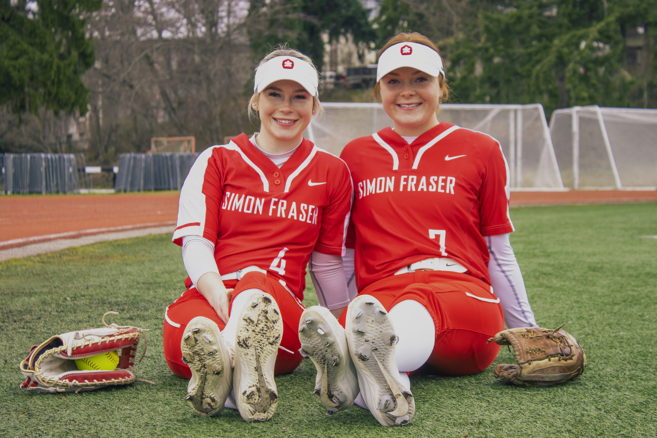 Georgia Ogg and Alex Ogg, two sisters on the SFU softball team, sit side by side with their legs out on the field, and a glove on either side of them.