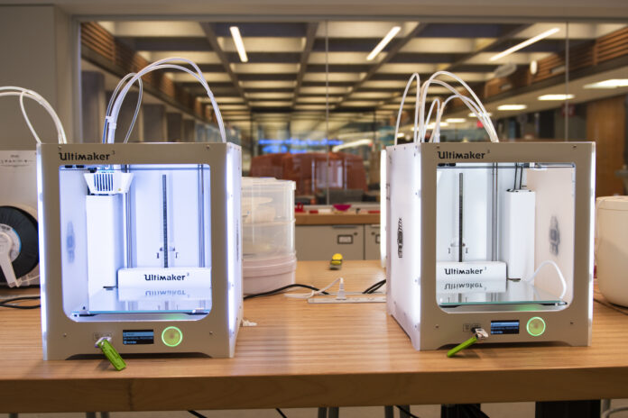 Two 3D printers on a worktable, backlit by fluorescent lighting