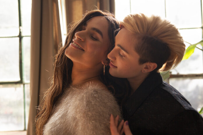 A couple are leaning into each other. Both appear somewhat femme, and look like they’re enjoying themselves.