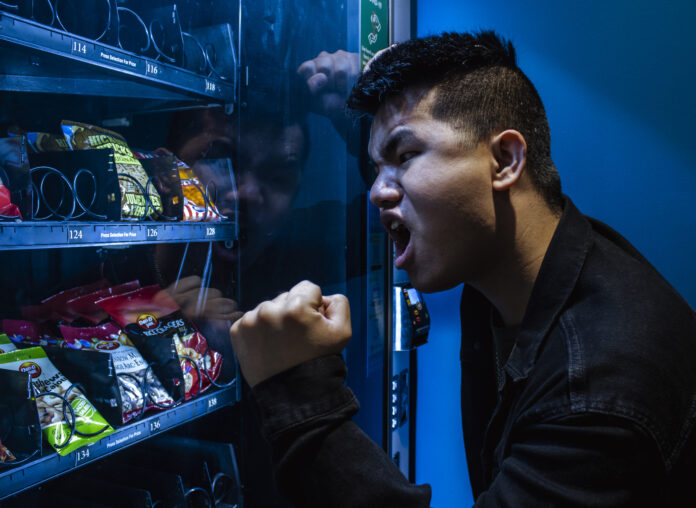 A man is yelling at a vending machine. The heel of his fist is against the glass, and he is leaning against the side of the machine. On his face is a look of pure outrage.