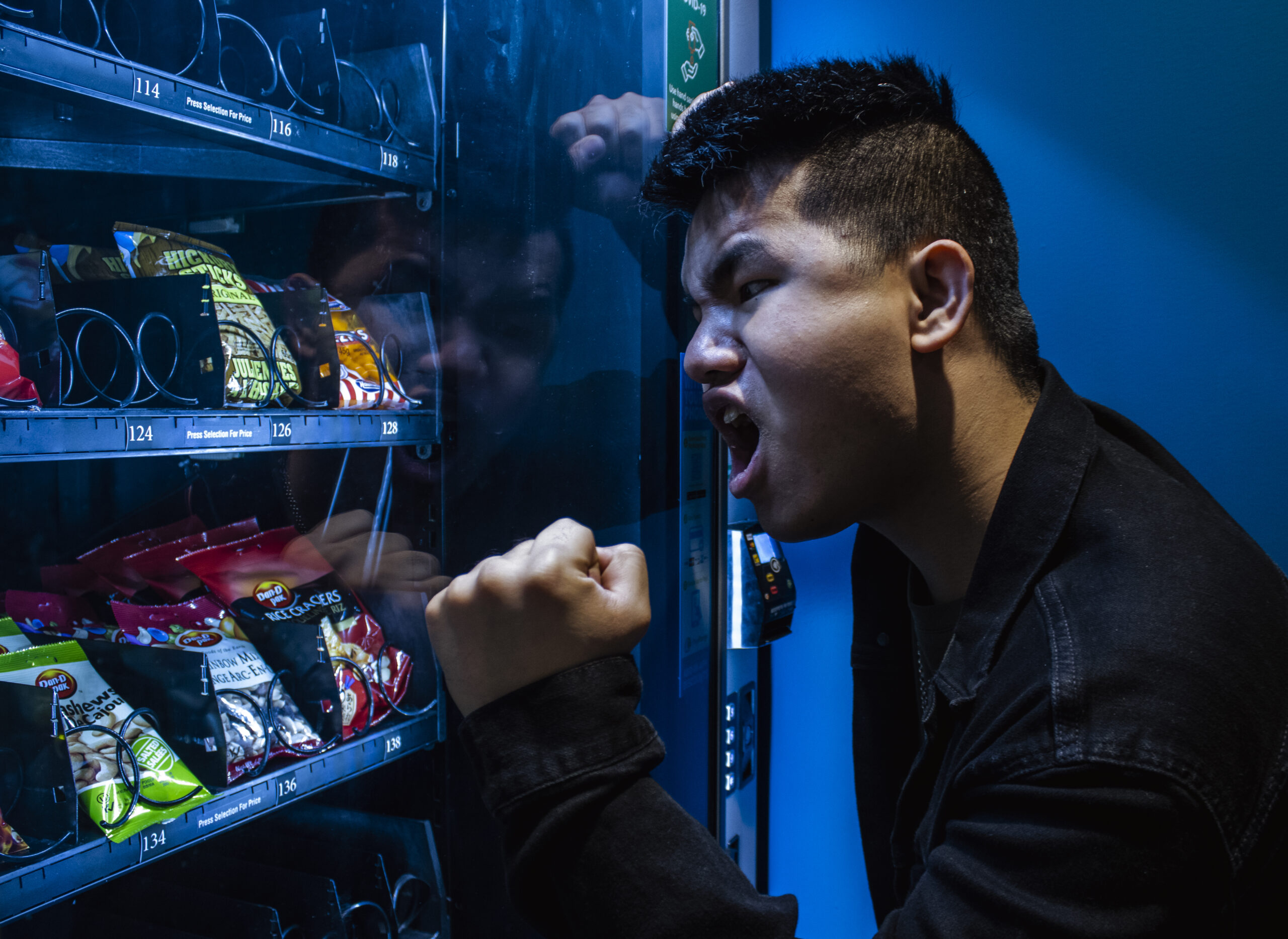 A man is yelling at a vending machine. The heel of his fist is against the glass, and he is leaning against the side of the machine. On his face is a look of pure outrage.