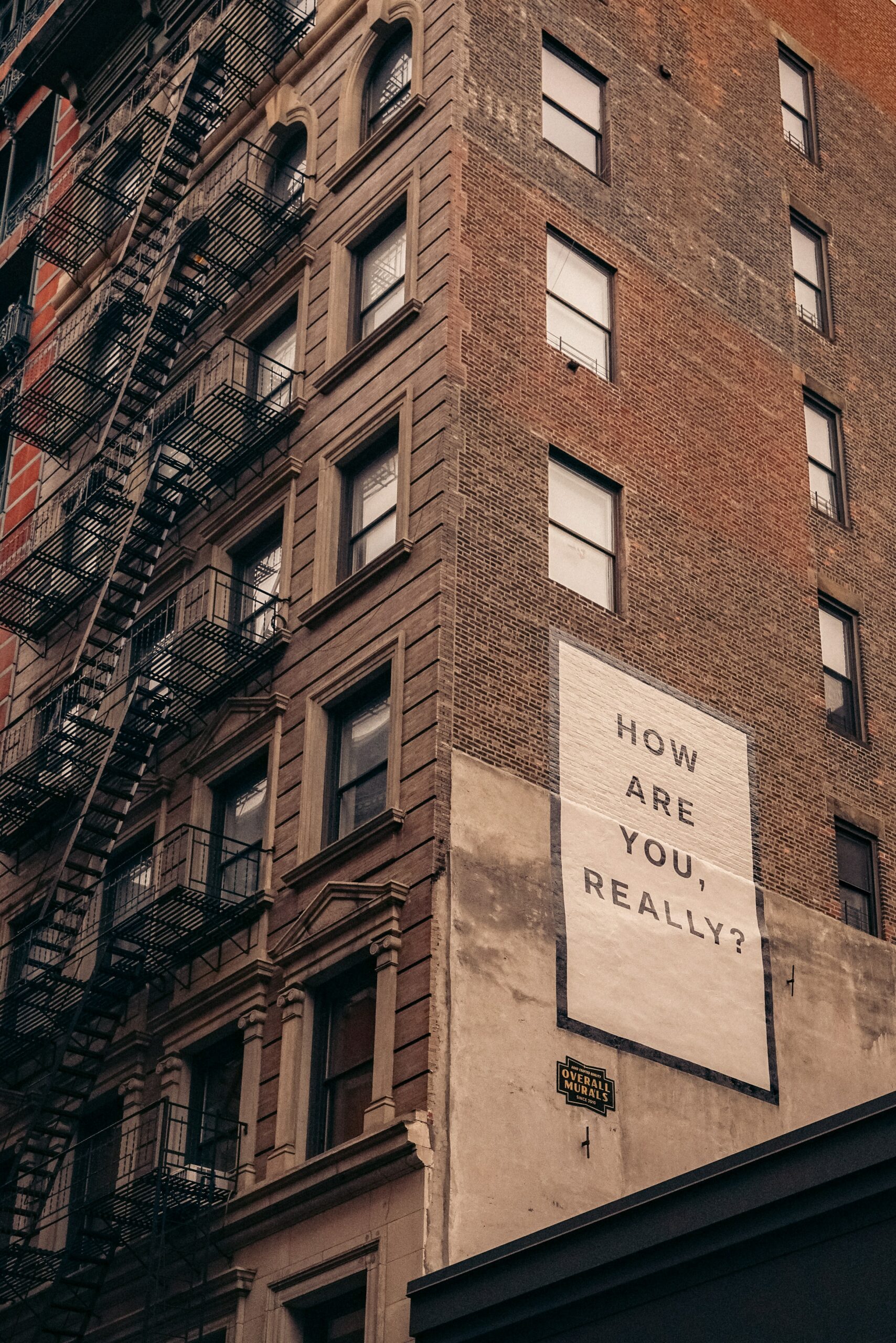 A message is painted on the side of a brick building. It reads “How are you, really?”